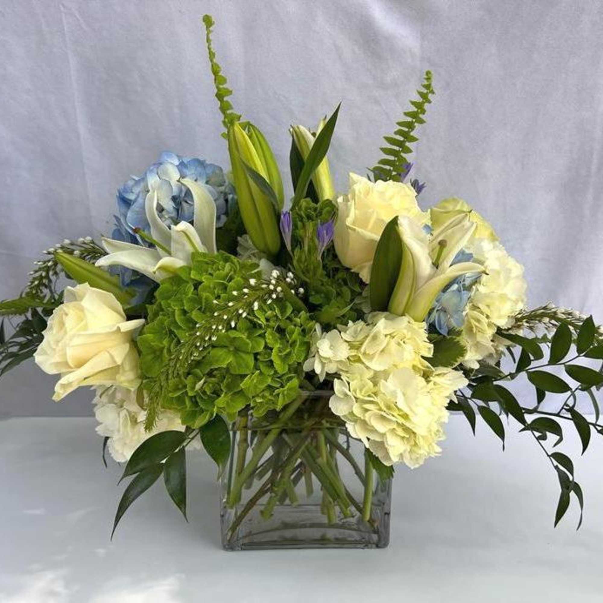 Arrangement of white roses, lilies, and green and blue hydrangeas in a clear glass cube vase