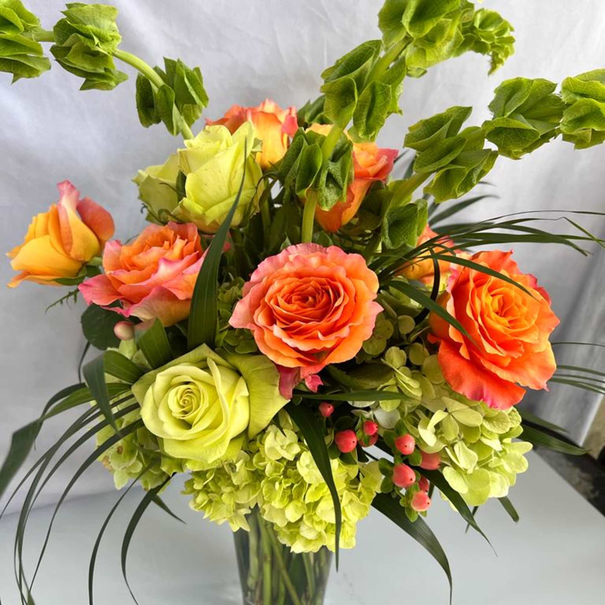 Tall arrangement of orange and green roses, green hydrangeas, and bells of Ireland in a clear glass vase