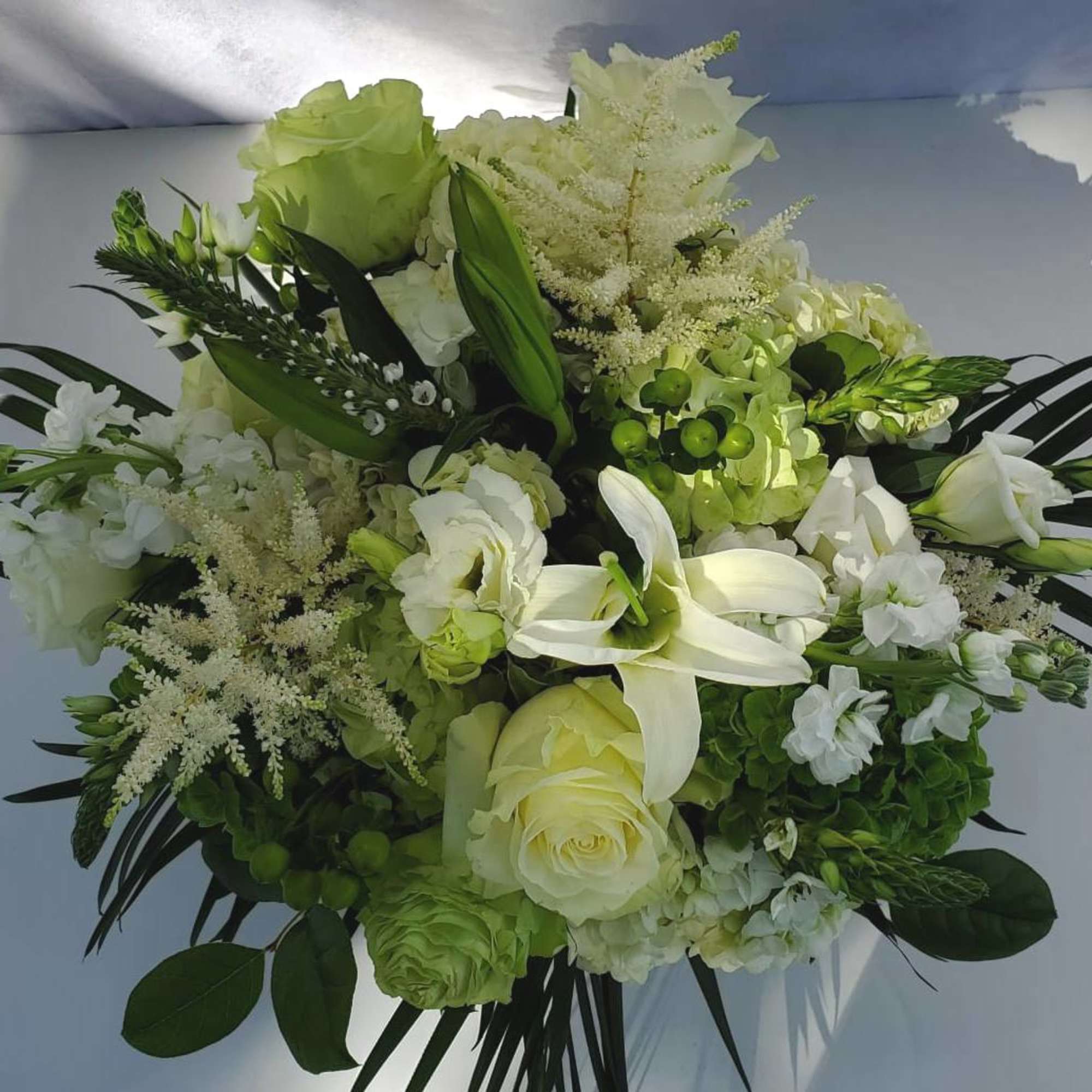 Round arrangement of white lilies, cream roses, and green hydrangeas with dark palm fronds
