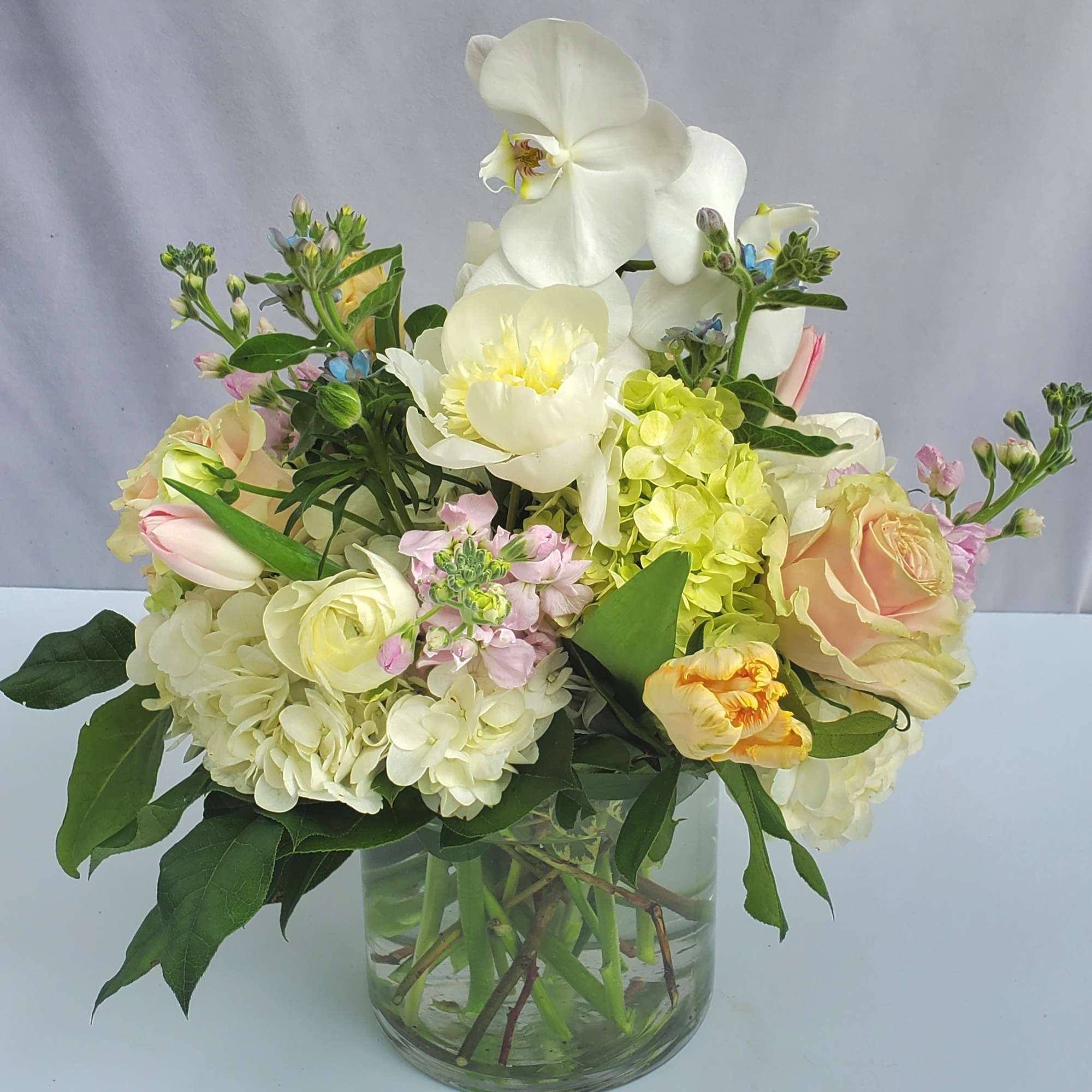 Pastel arrangement of orchids, roses, tulips, and hydrangeas in a clear glass cylinder vase