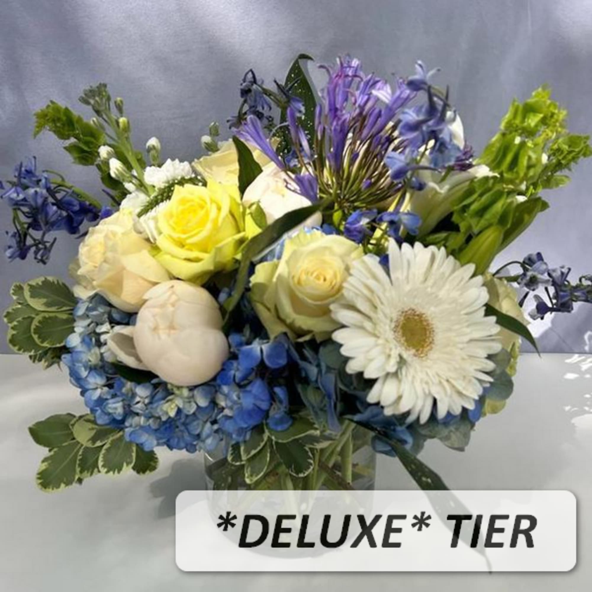 Deluxe bouquet of yellow roses, blue hydrangeas, and white gerbera daisies in a clear glass vase