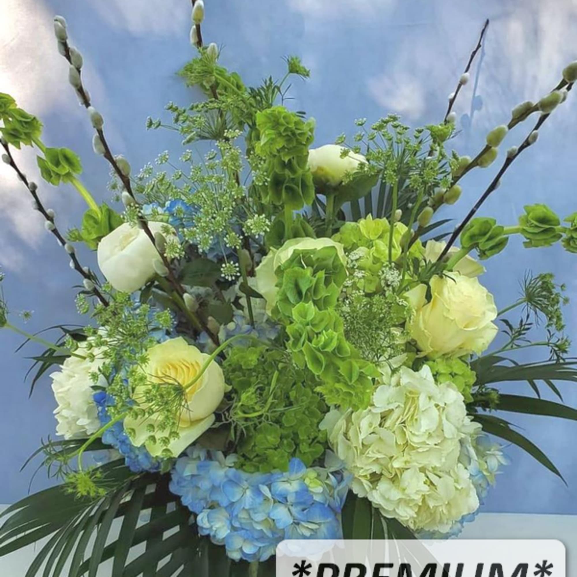 Tall arrangement of green bells of Ireland, white roses, and blue hydrangeas in a clear glass vase