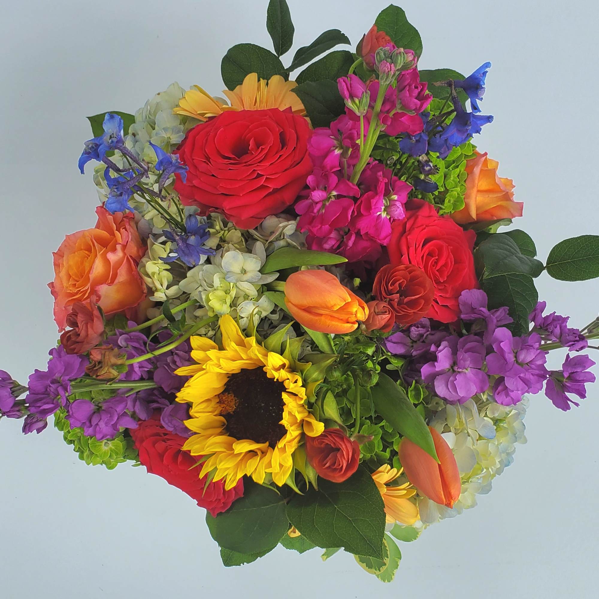 Bright mixed arrangement with red roses, yellow sunflower, orange tulips, hydrangea and purple flowers.