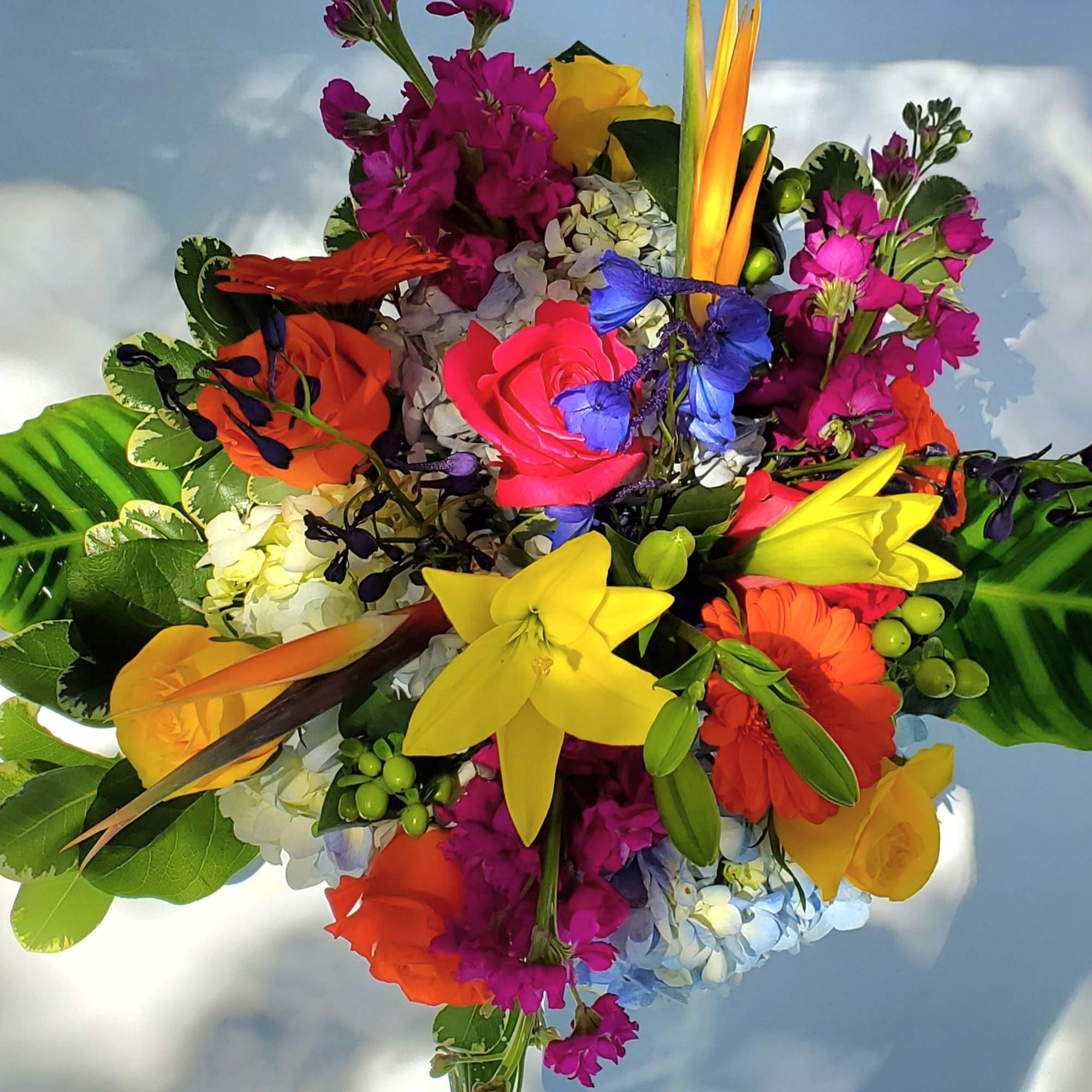 Arrangement of bright tropical flowers with yellow lilies, roses, and gerbera daisies, accented by bold striped tropical leaves.