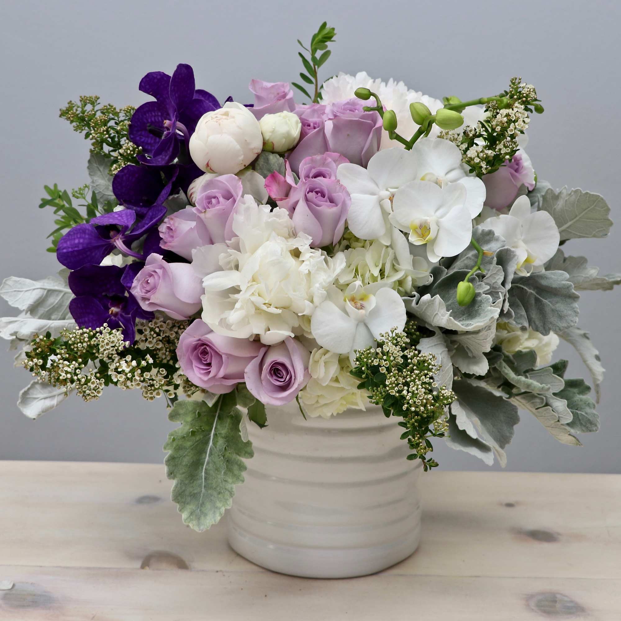Low white vase filled with lavender roses, white hydrangeas, and orchids with purple accents
