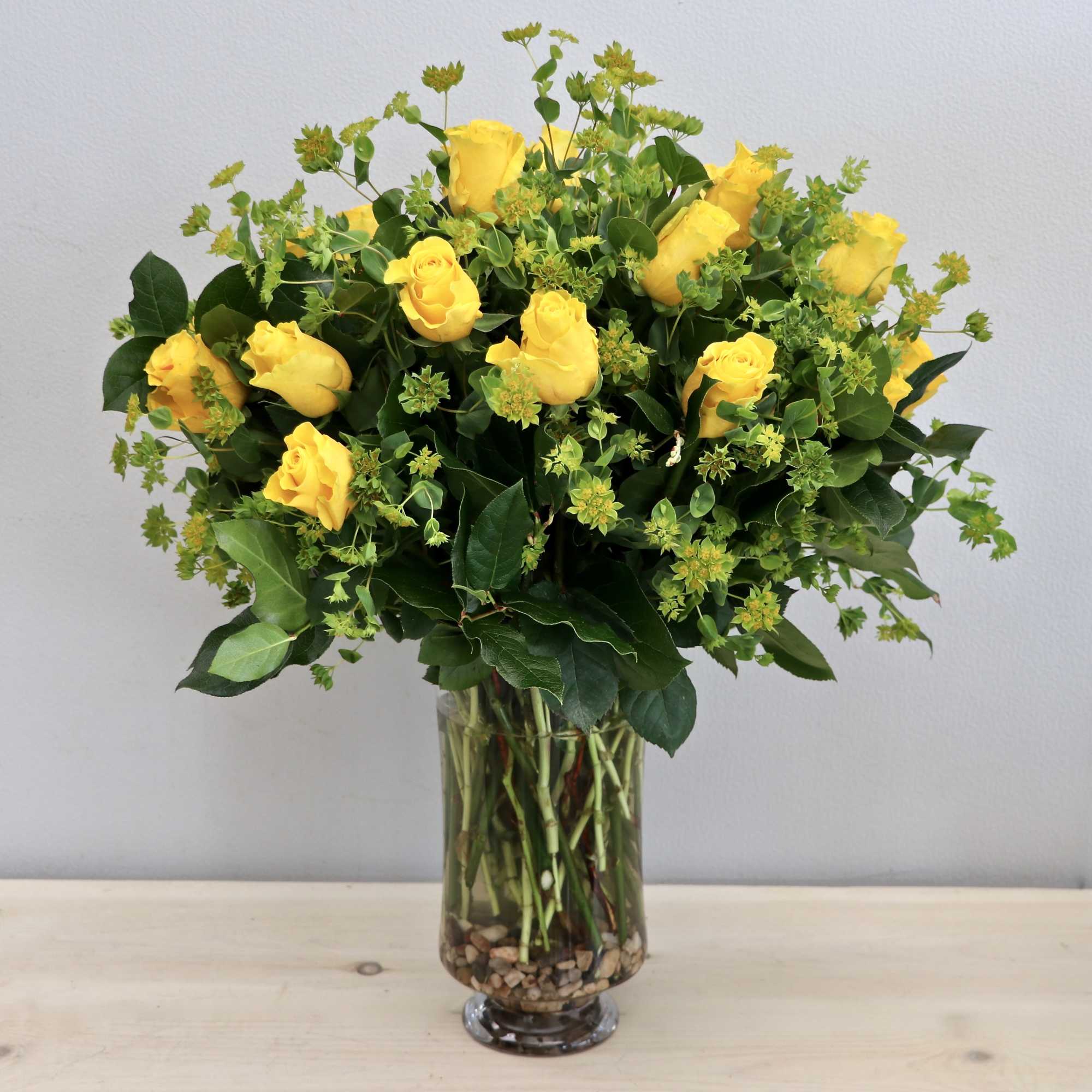 Tall arrangement of yellow roses with filler blooms in a clear glass vase with stones
