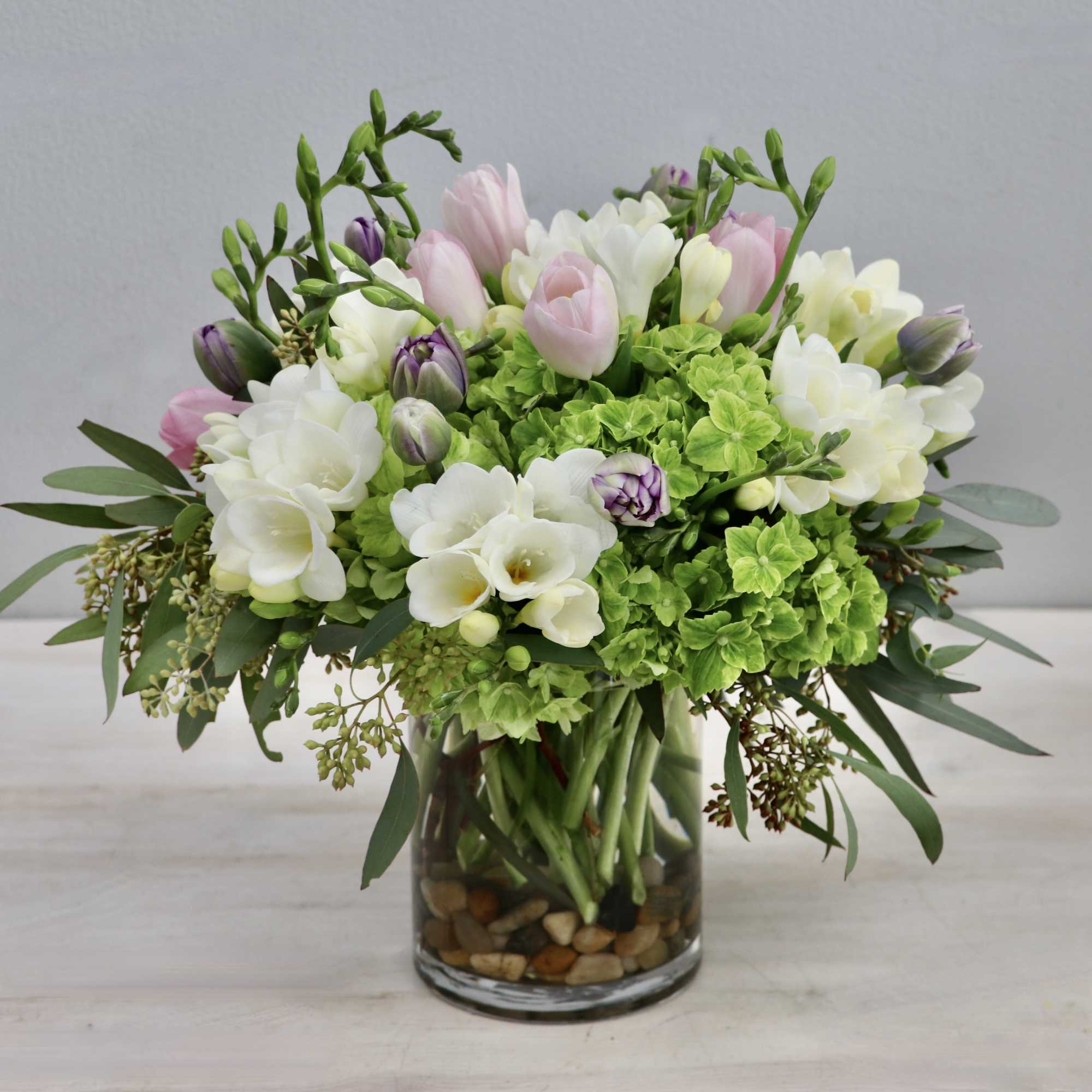 Compact arrangement of pink tulips, white freesias, and green hydrangeas in a clear glass vase with stones.