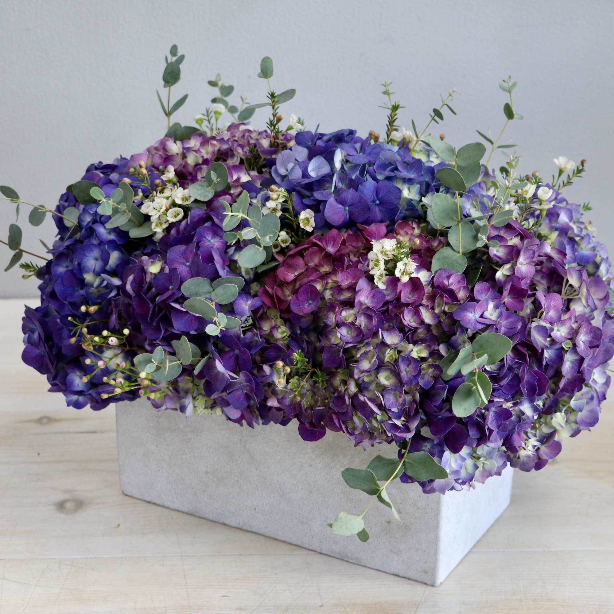 Low arrangement of purple and lavender hydrangeas with small white blooms in a rectangular light gray container