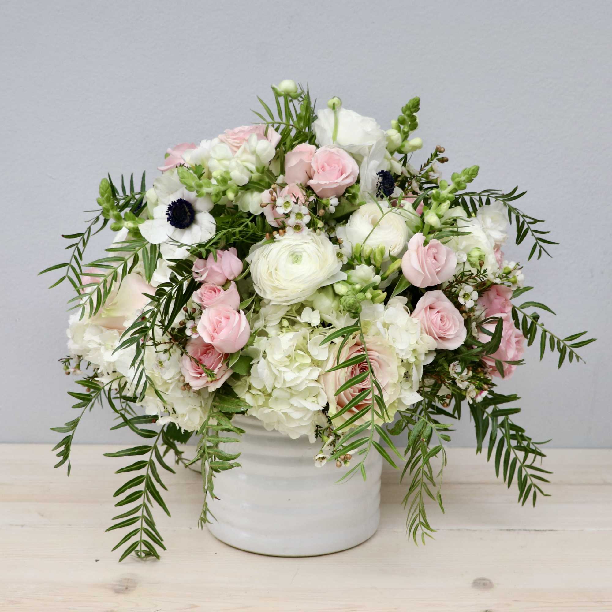 White and blush flower arrangement with trailing greenery in a white ceramic vase.