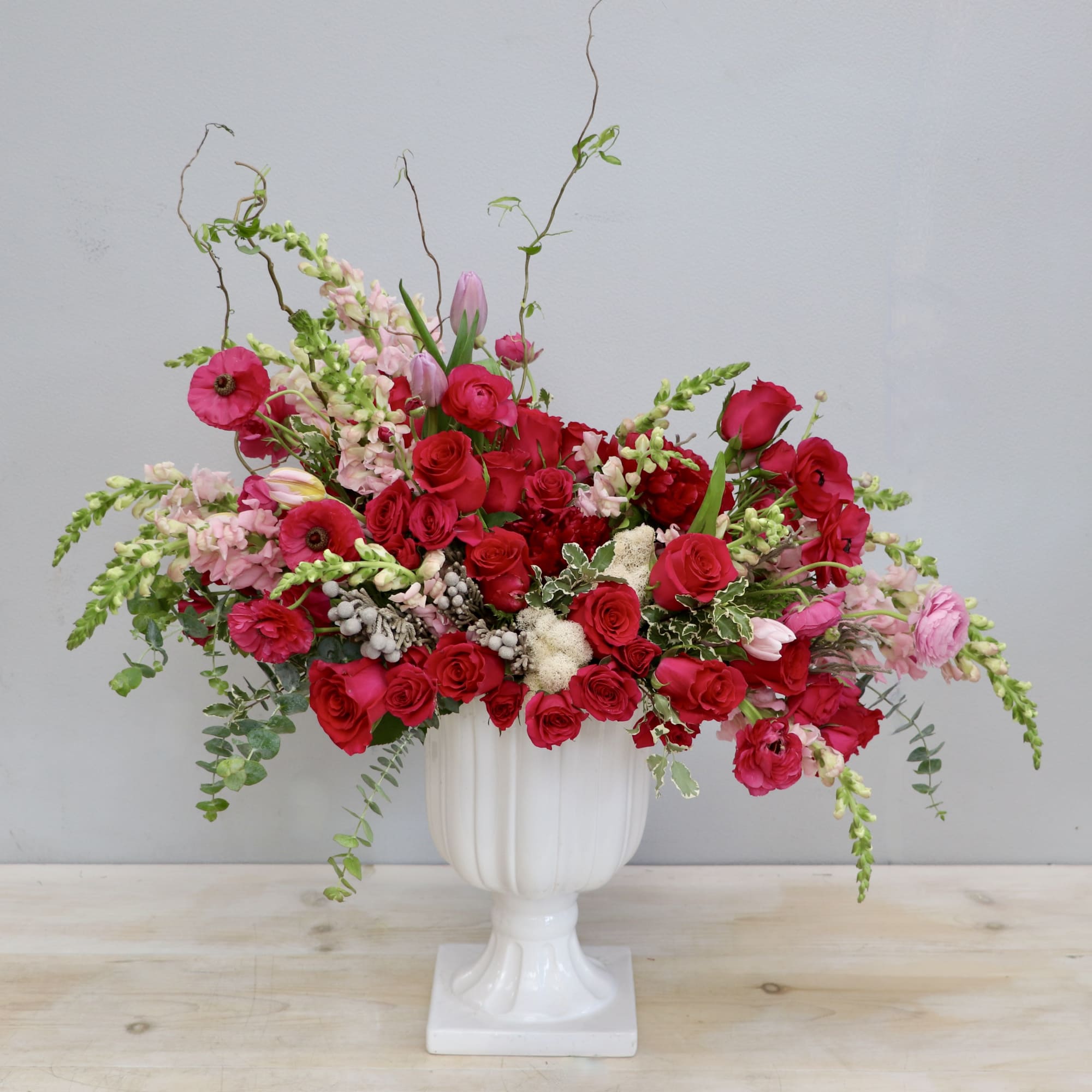 Lush red and pink flower arrangement in a white pedestal vase with trailing blooms and curly branches