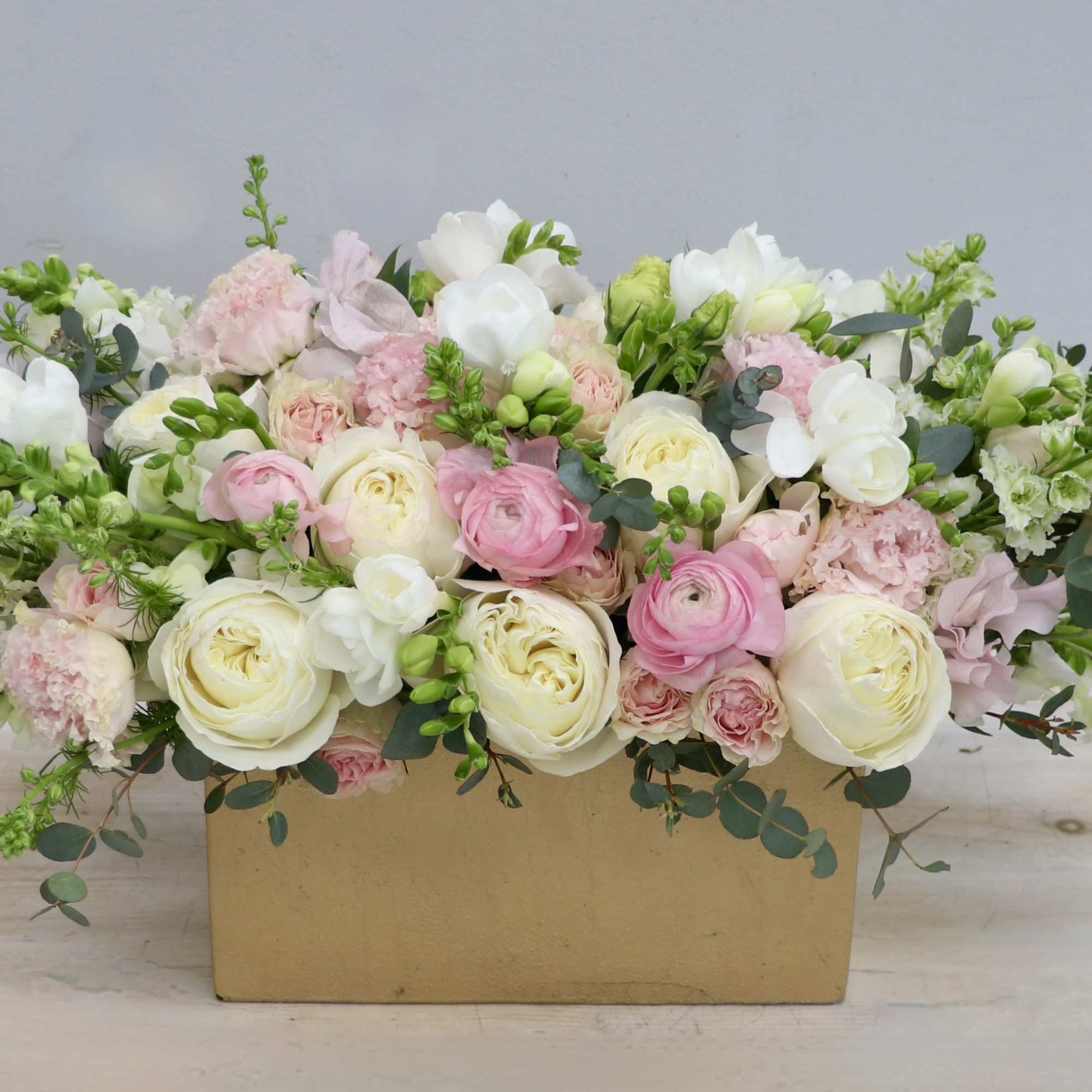 Low rectangular arrangement of white and pink flowers with trailing green accents in a gold container