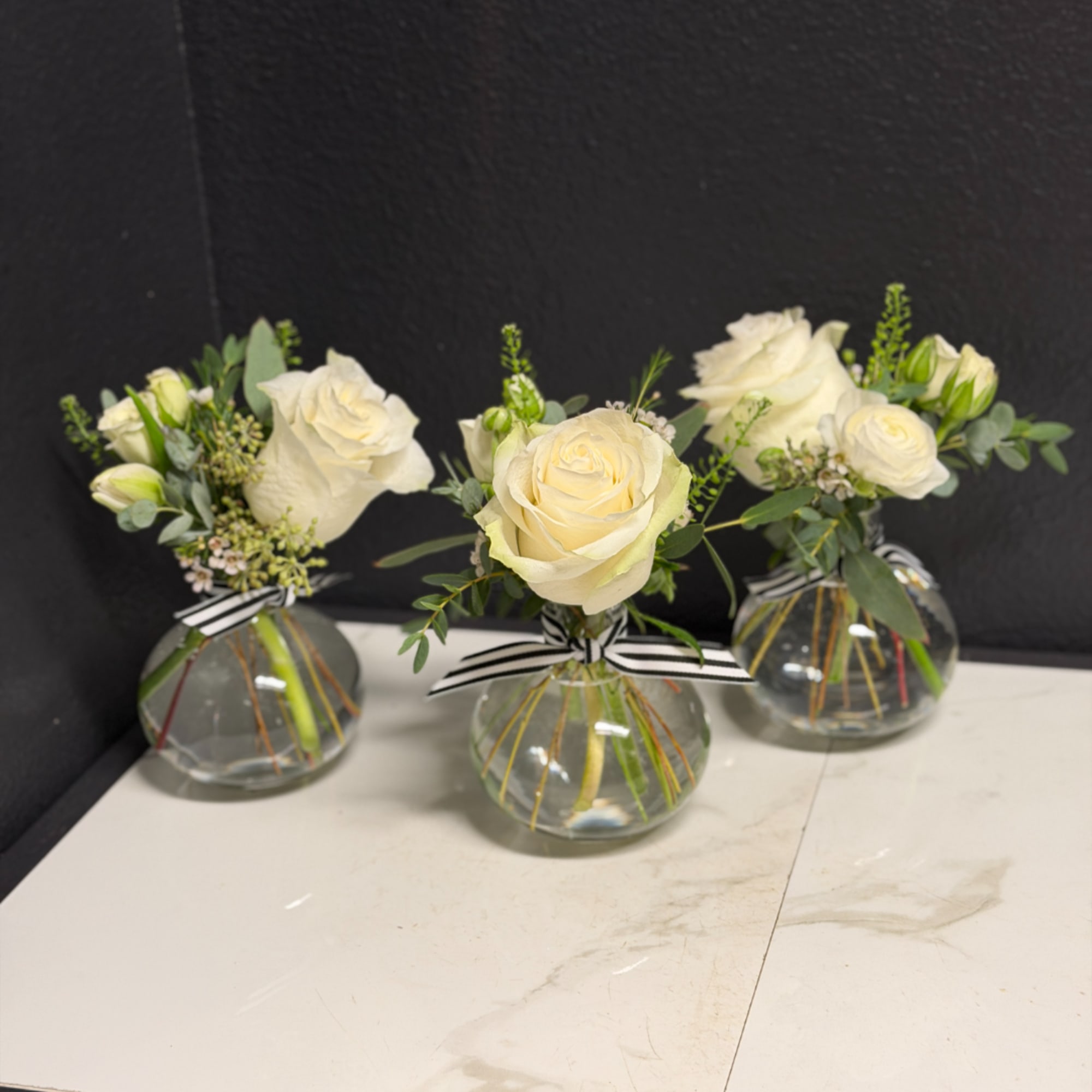 Three small white rose arrangements in round glass vases with striped ribbons.
