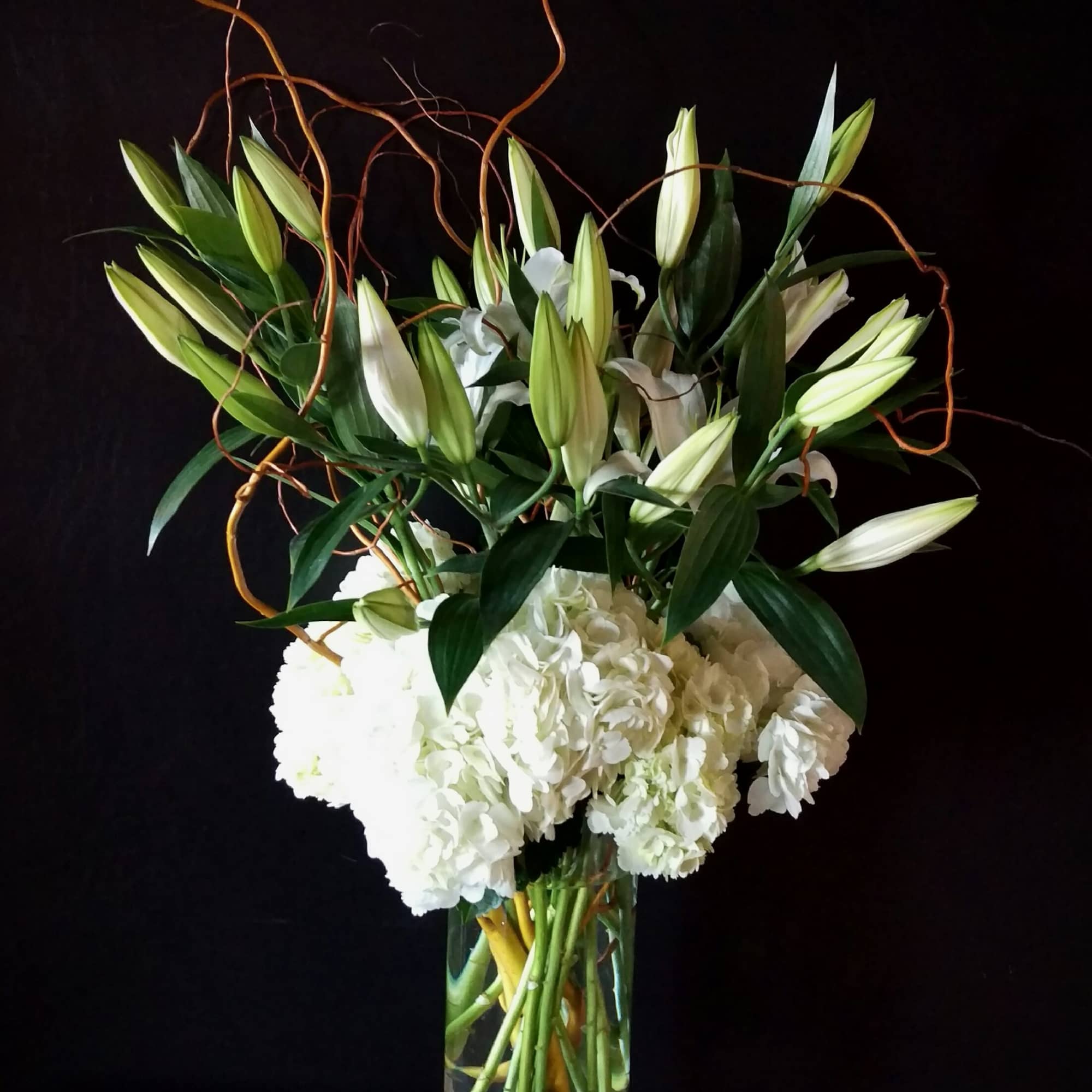 White lilies and hydrangeas in a tall glass vase with curly branches