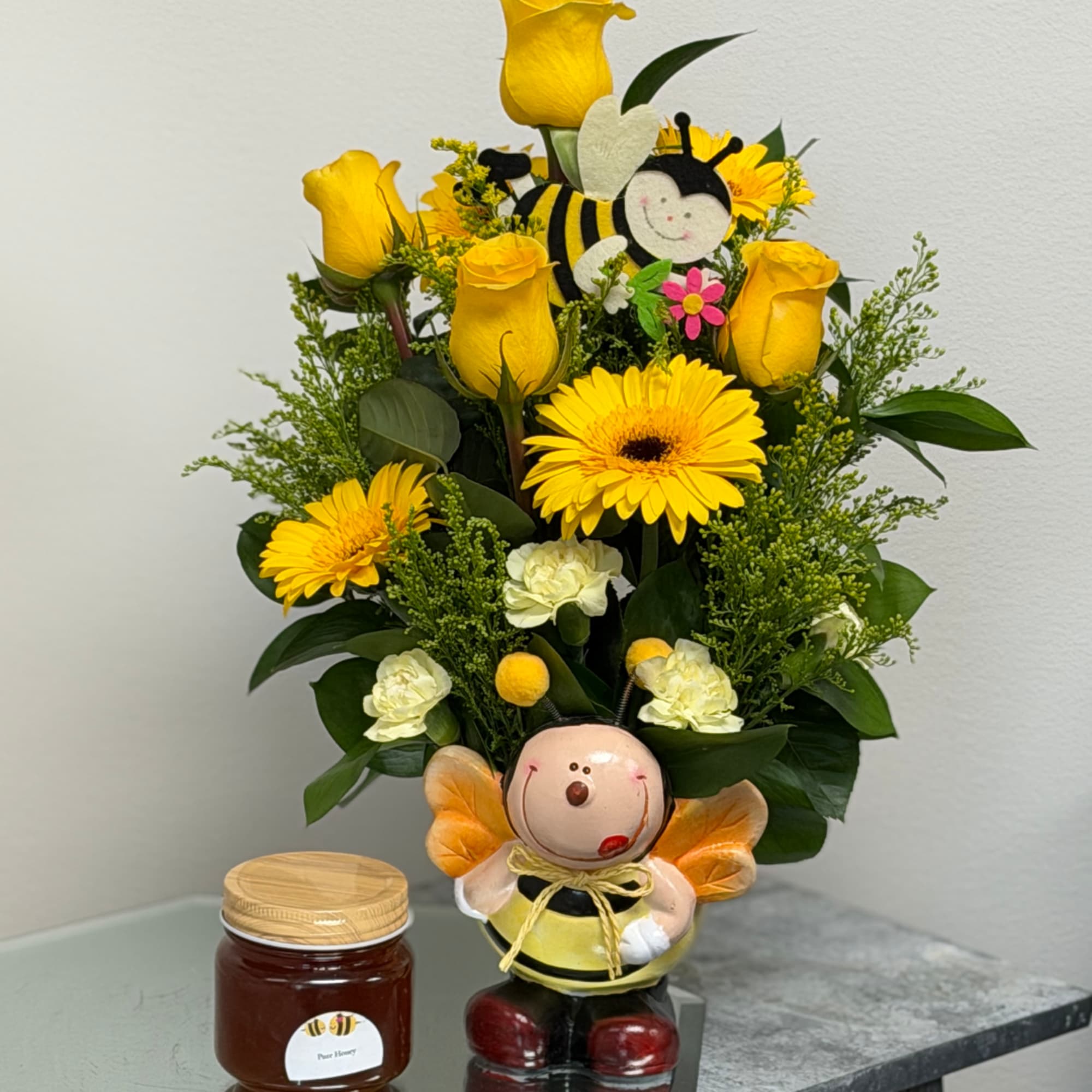 Yellow roses and gerbera daisies in a bee-themed vase with a plush bee topper.