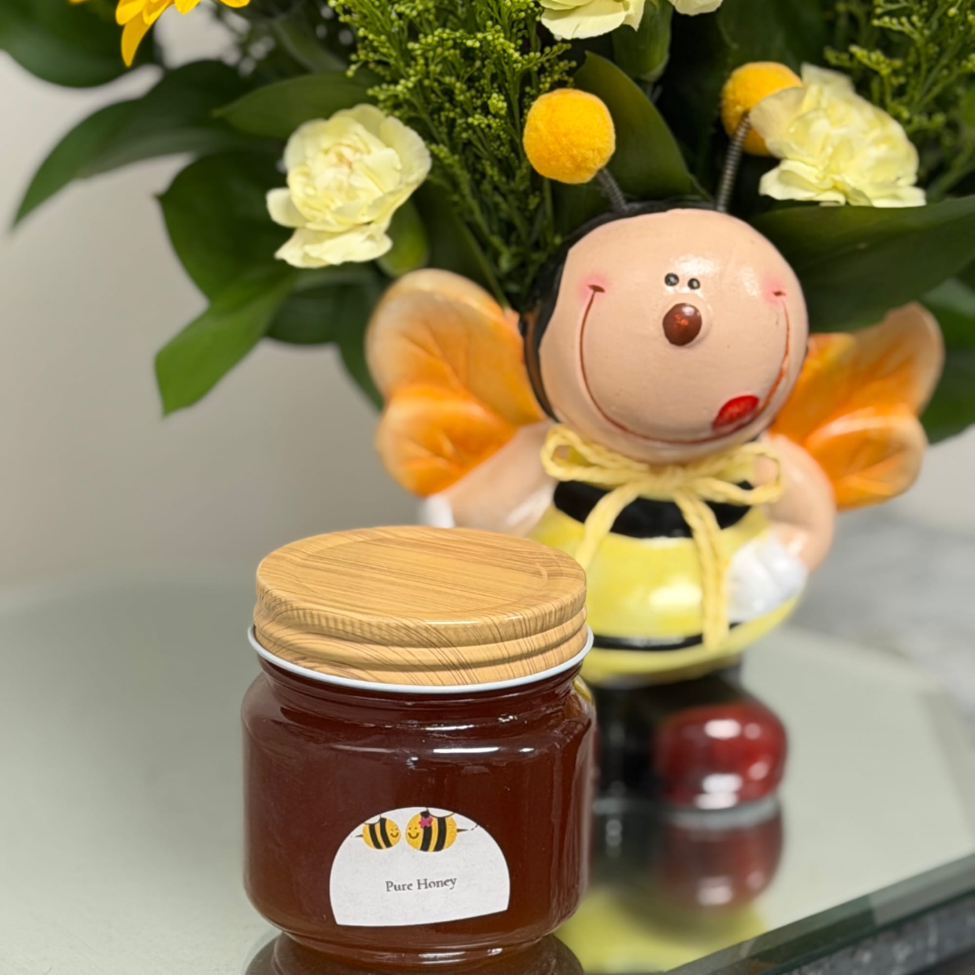 Jar of honey with a wooden lid in front of a bee-themed figurine and flowers.