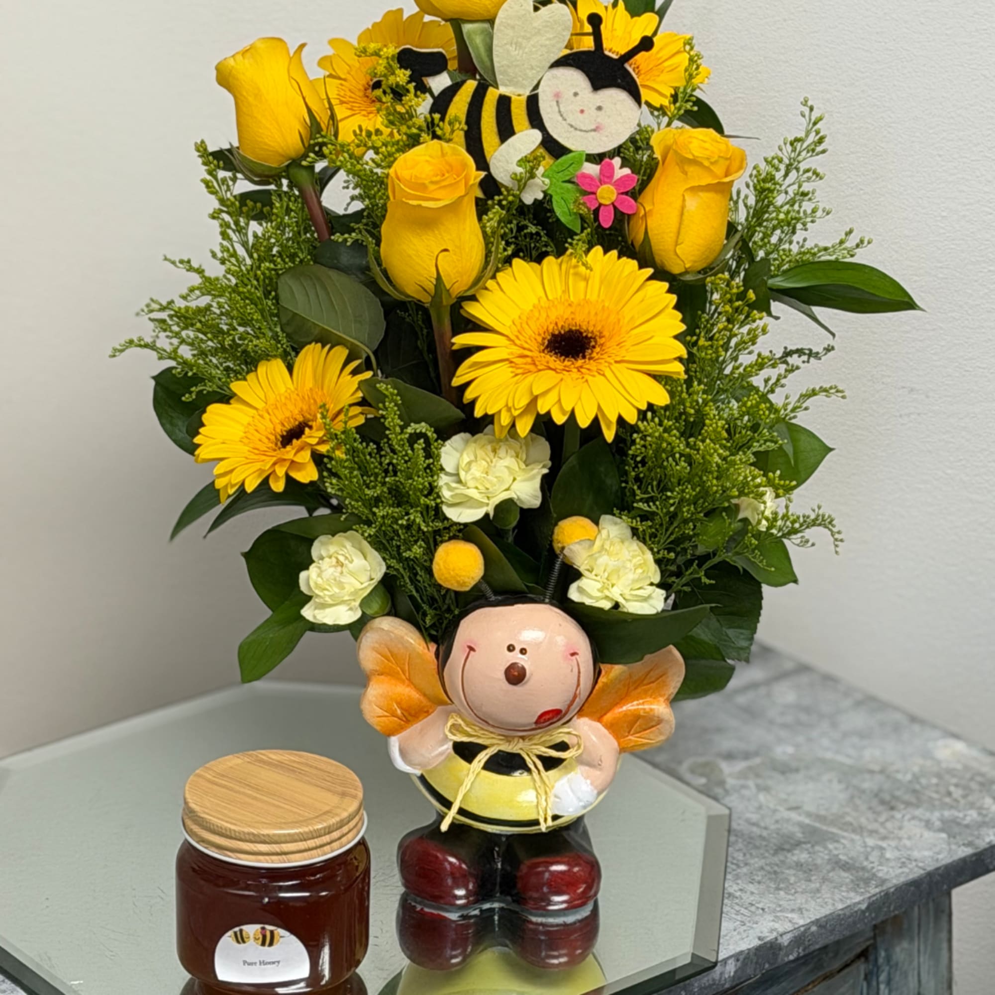 Yellow roses and daisies arranged in a bee-themed vase with a small honey jar beside it.