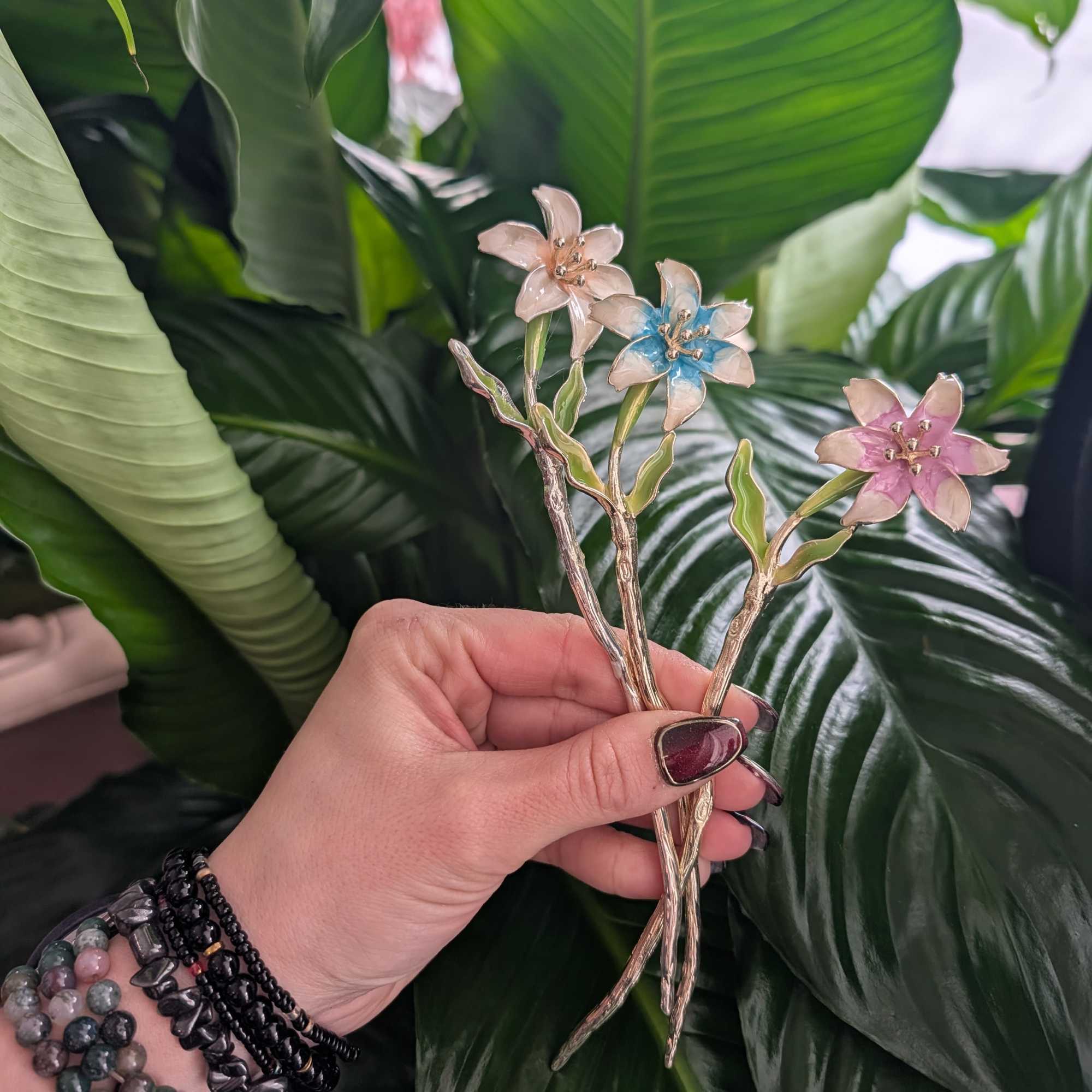 Hand holding three small enamel flower pins against large green leaves
