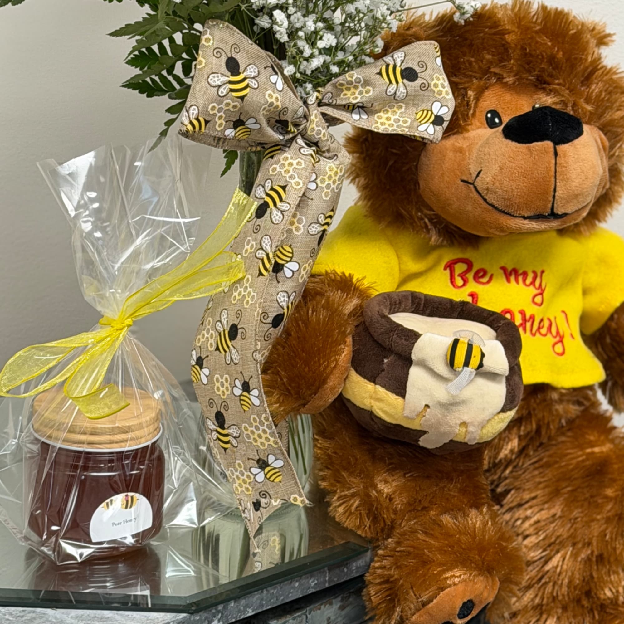 Teddy bear with a bee-themed shirt beside a wrapped floral gift and honey jar