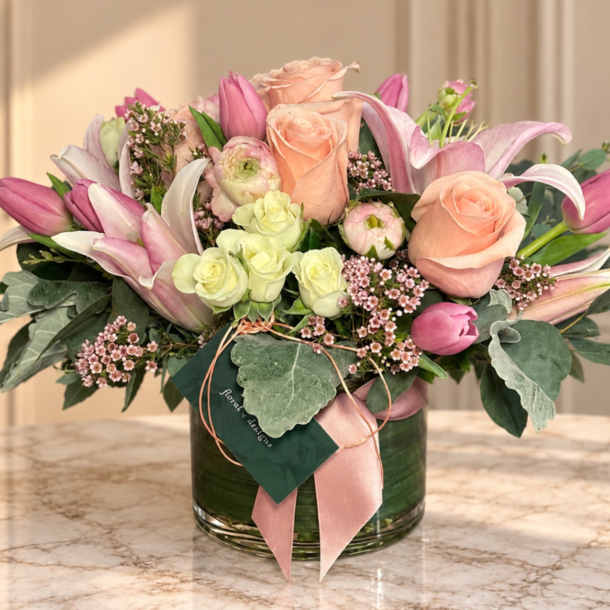 Pink lilies and peach roses arranged in a glass vase with ribbon.