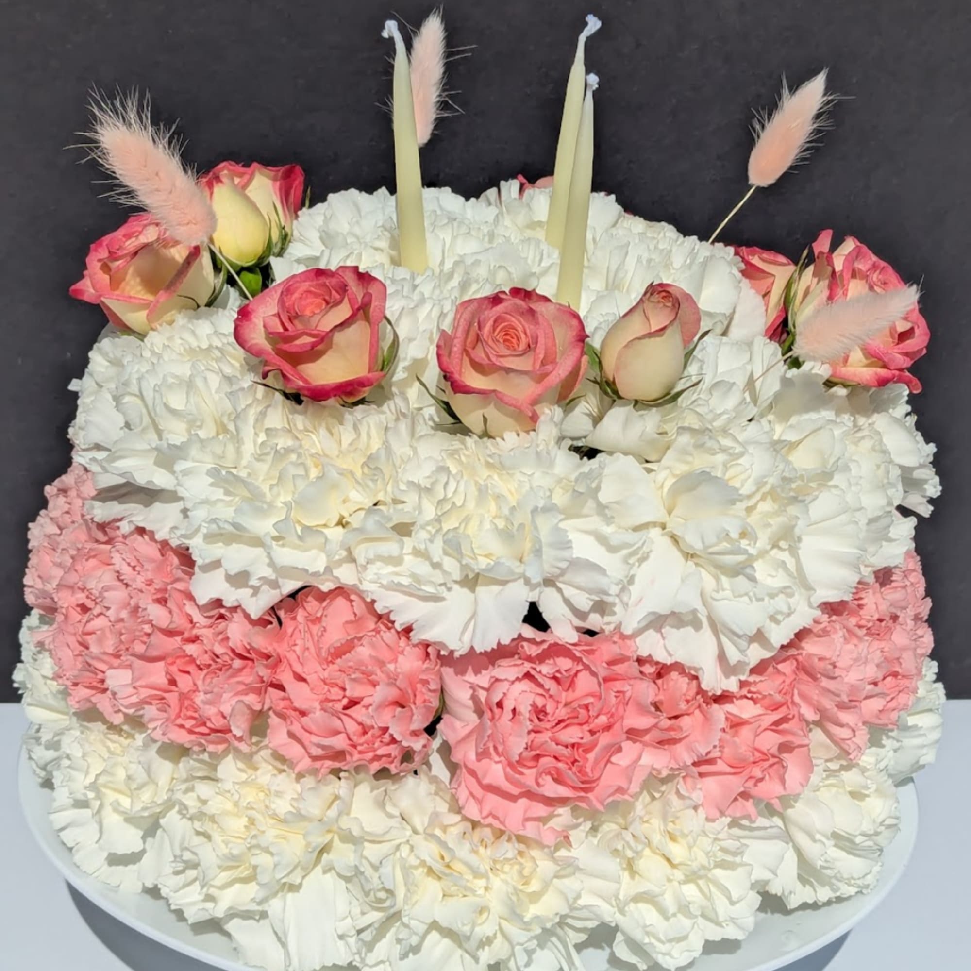 Pink roses and white carnations arranged on a cake-shaped base with candles.