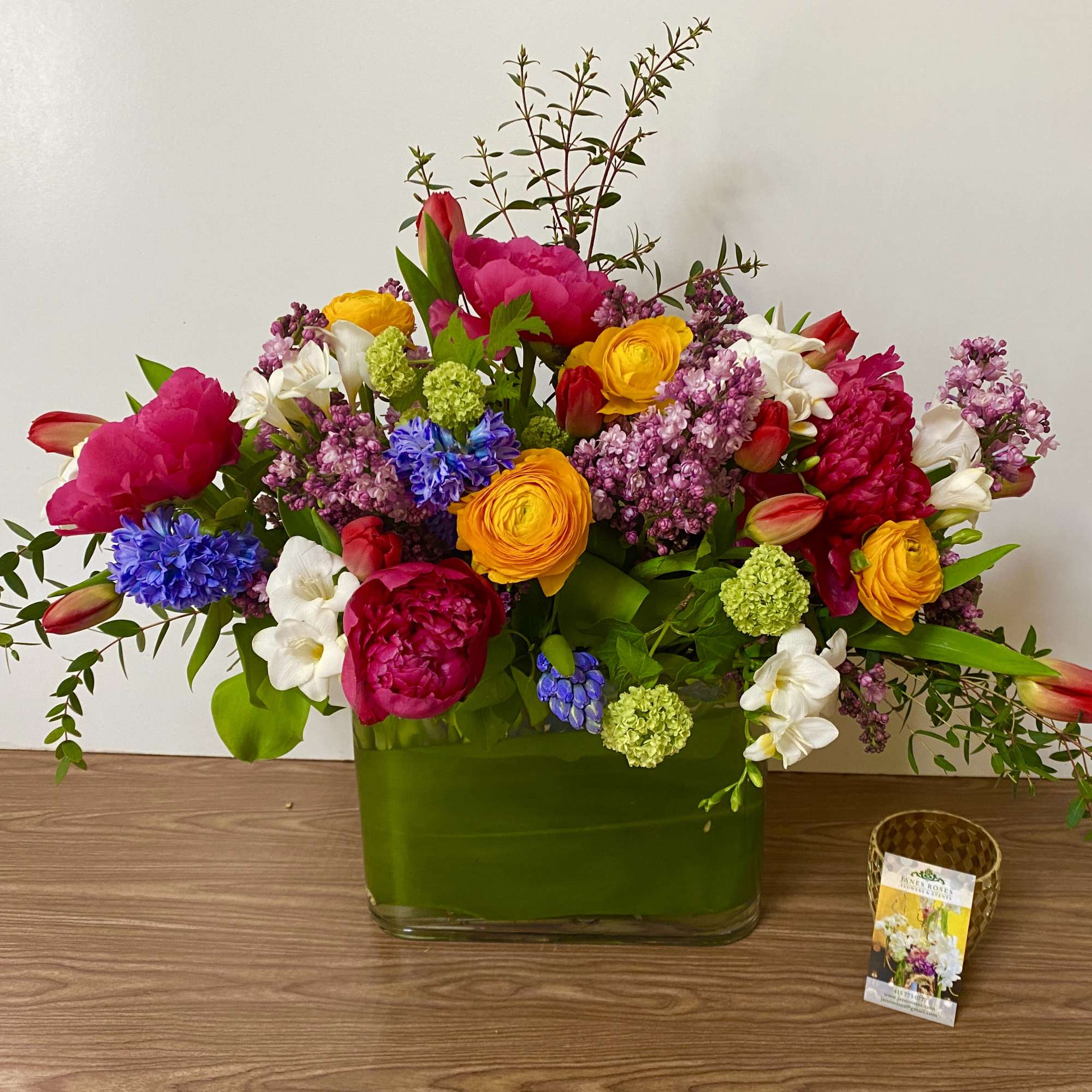 Bright mixed bouquet in a green glass vase with pink, yellow, white, and blue flowers.