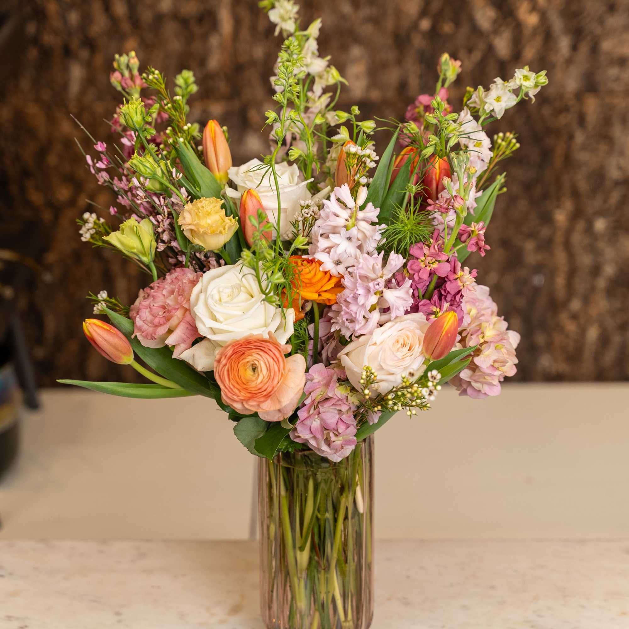 Tall glass vase of mixed roses, tulips, hydrangeas and spring blooms in soft pink, white and orange tones