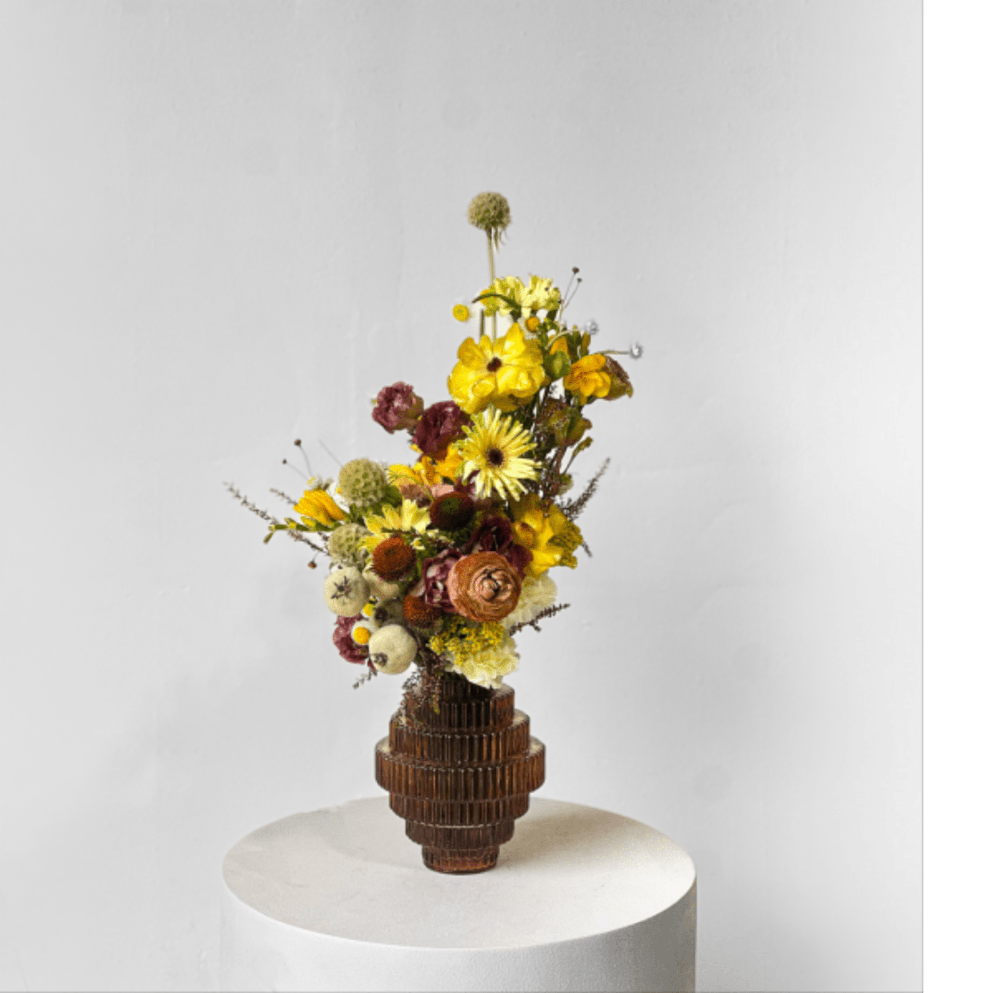 Tall mixed flower arrangement in a ribbed brown vase on a white pedestal