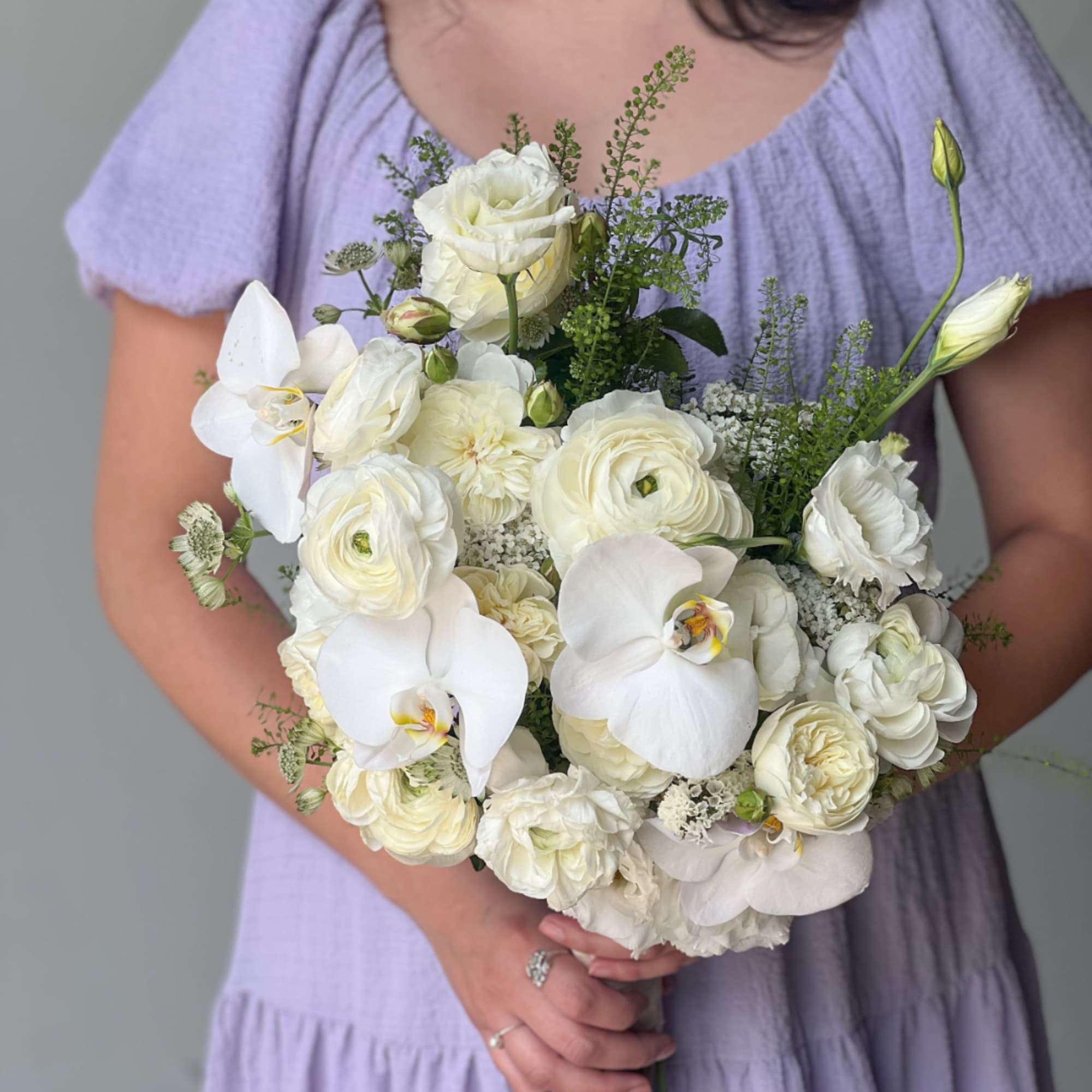 White bouquet with roses, orchids, and ranunculus held by a person in a lavender dress