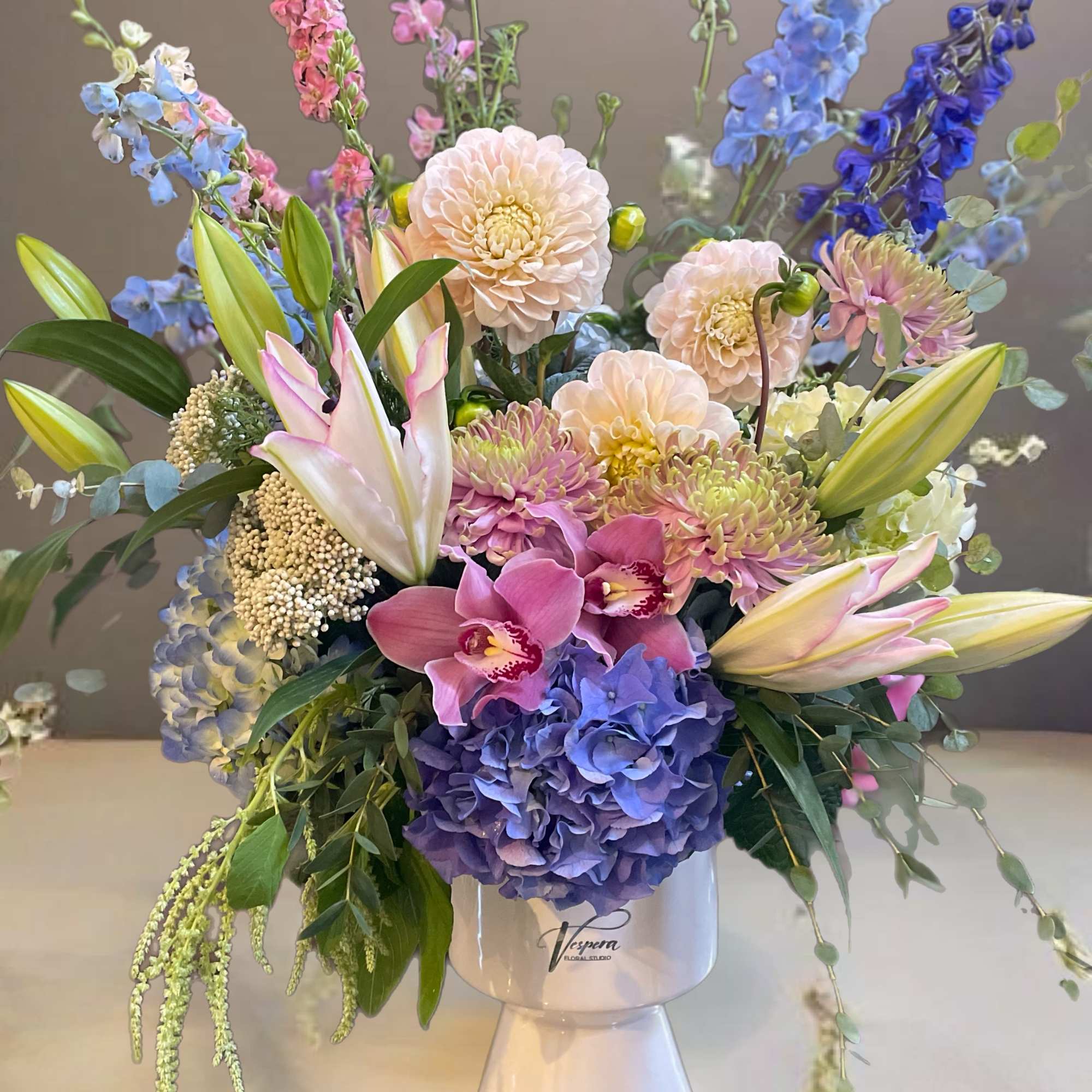 Colorful bouquet with lilies, hydrangeas, orchids, and tall blue flowers in a white vase