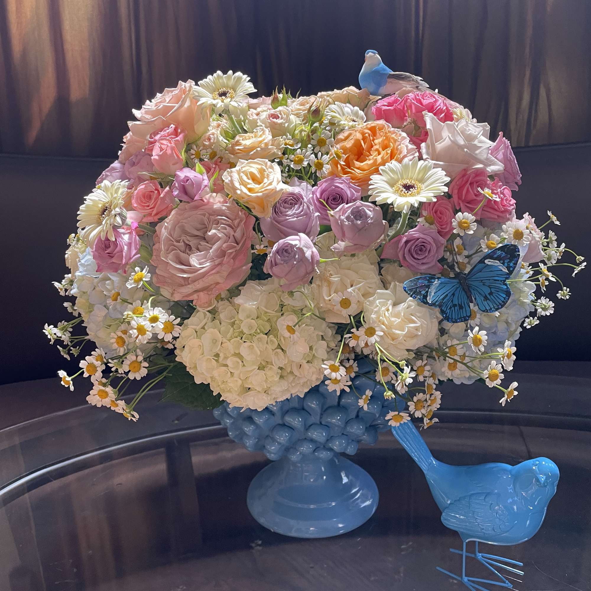 Pastel bouquet of roses, daisies, and hydrangeas in a blue vase with bird accents