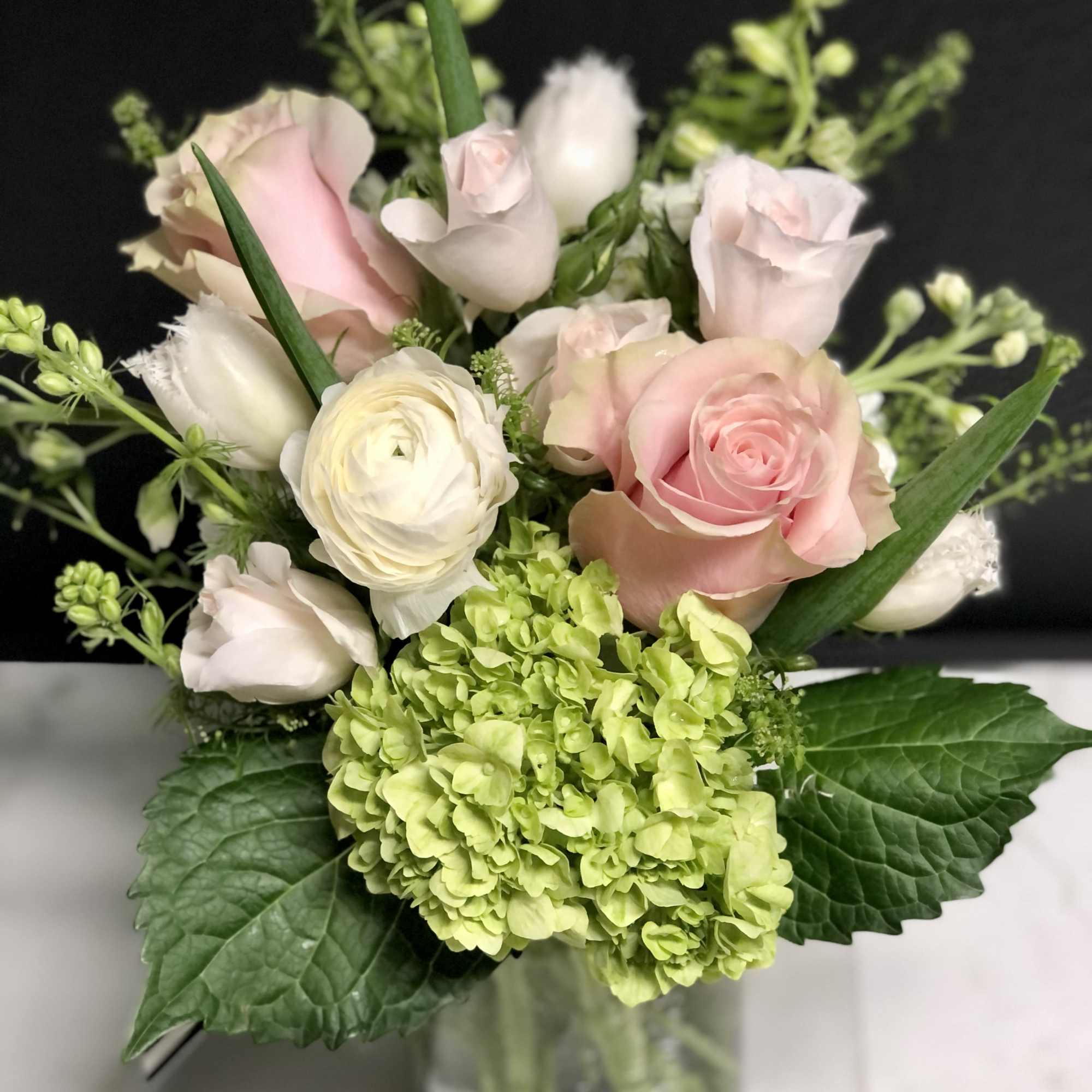 Short glass vase of pale pink roses, white blooms, and green hydrangea against a dark background