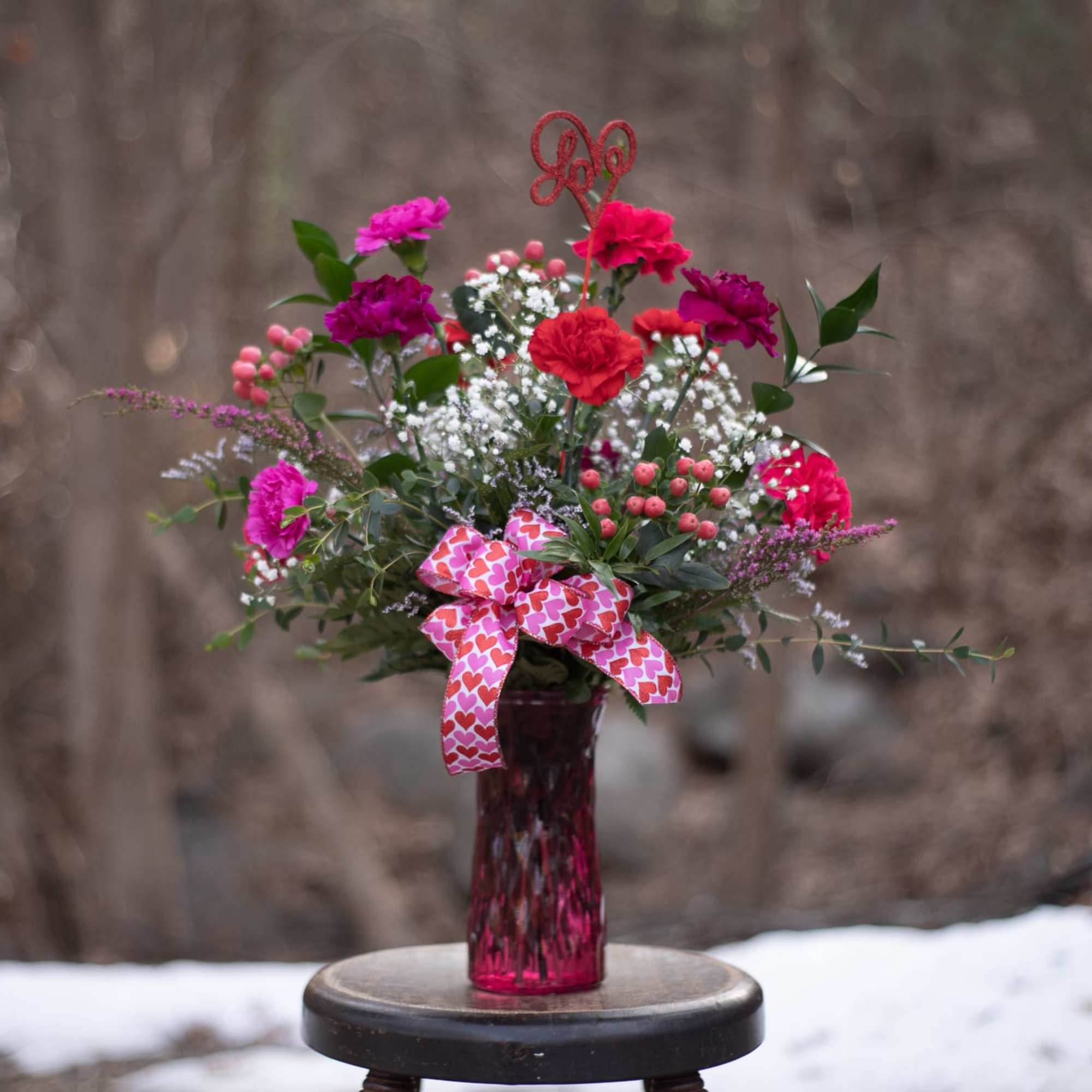 Pink and red flower arrangement in a red vase with a heart ribbon