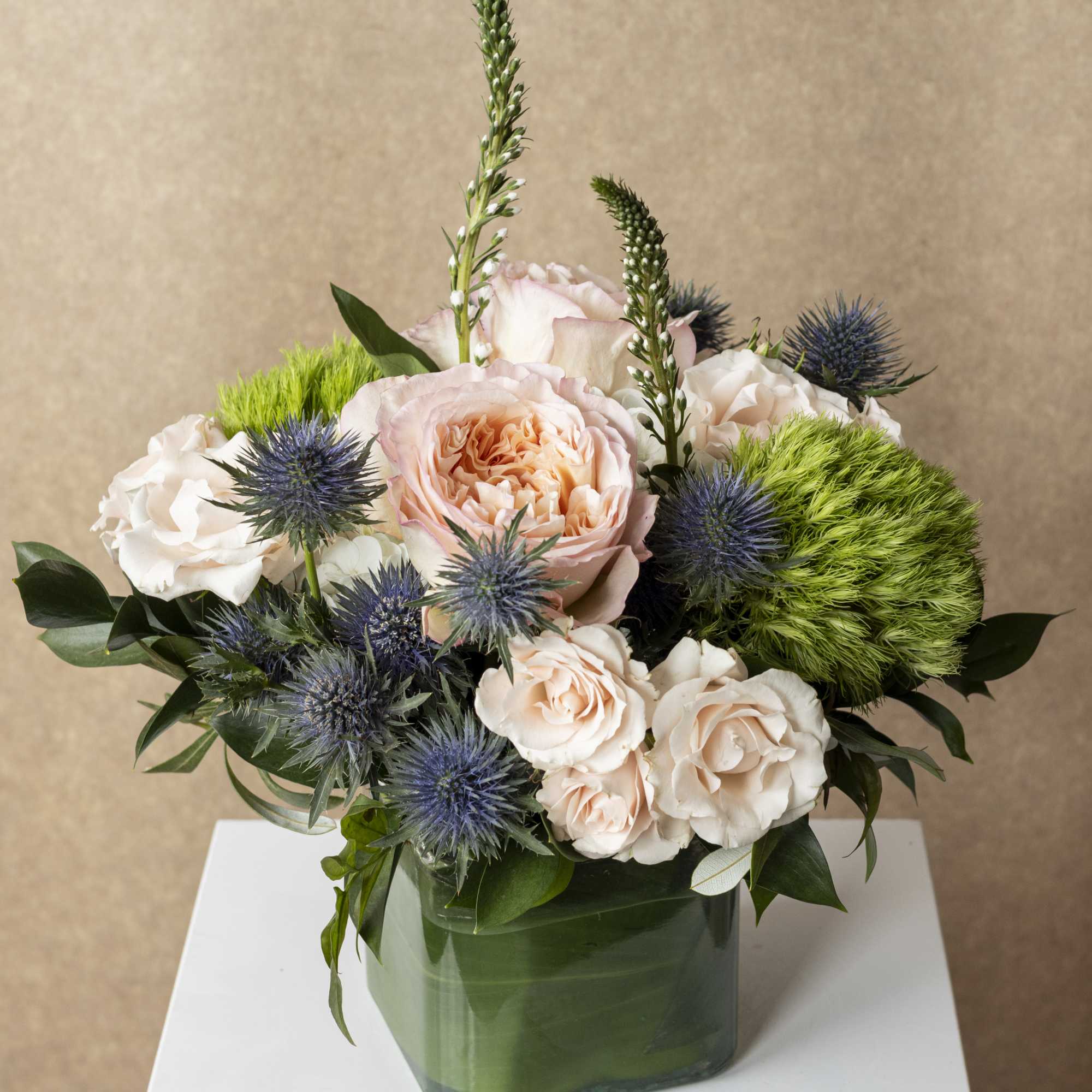 Peach and white roses in a green glass vase with blue thistle and green pom-pom flowers