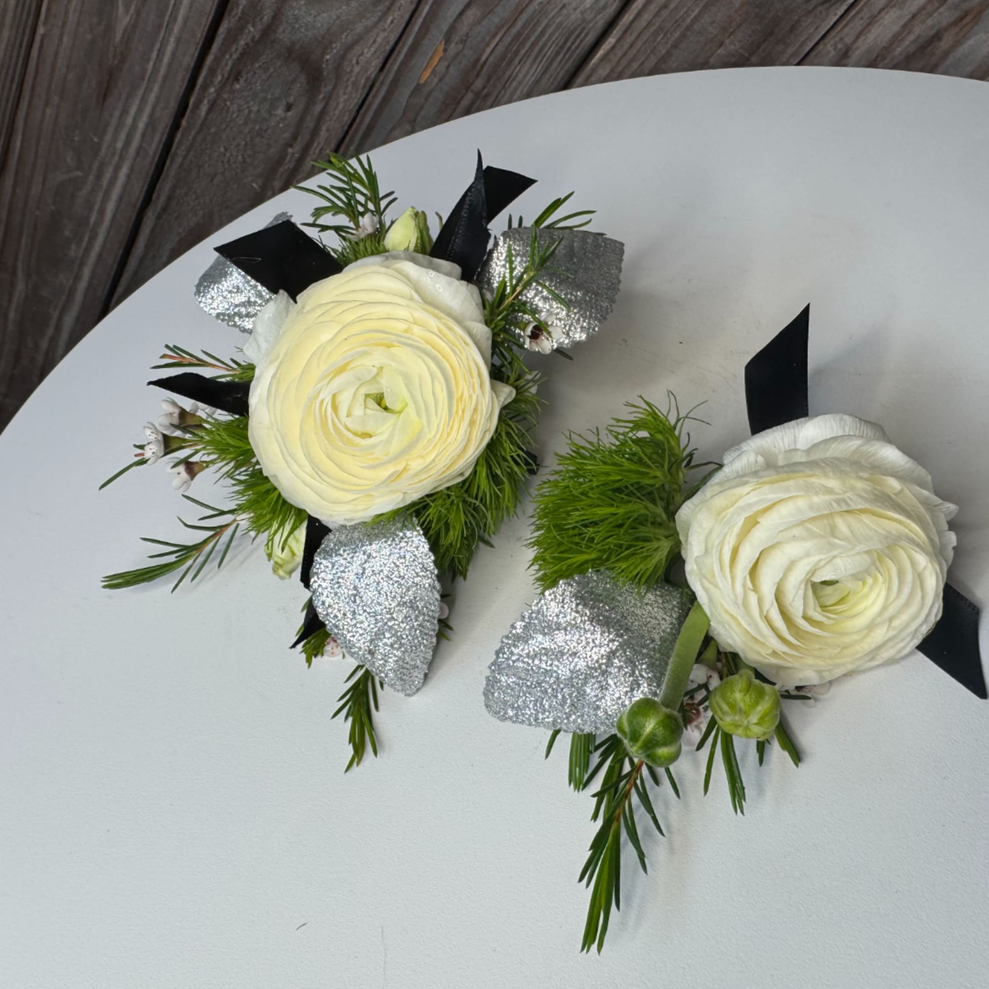 Two white ranunculus boutonnieres with silver accents and black ribbon