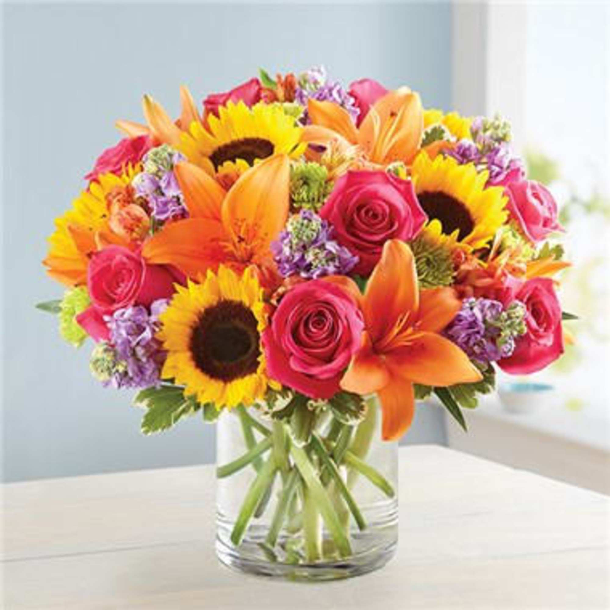 Bright mixed bouquet of sunflowers, roses, and lilies in a clear glass vase