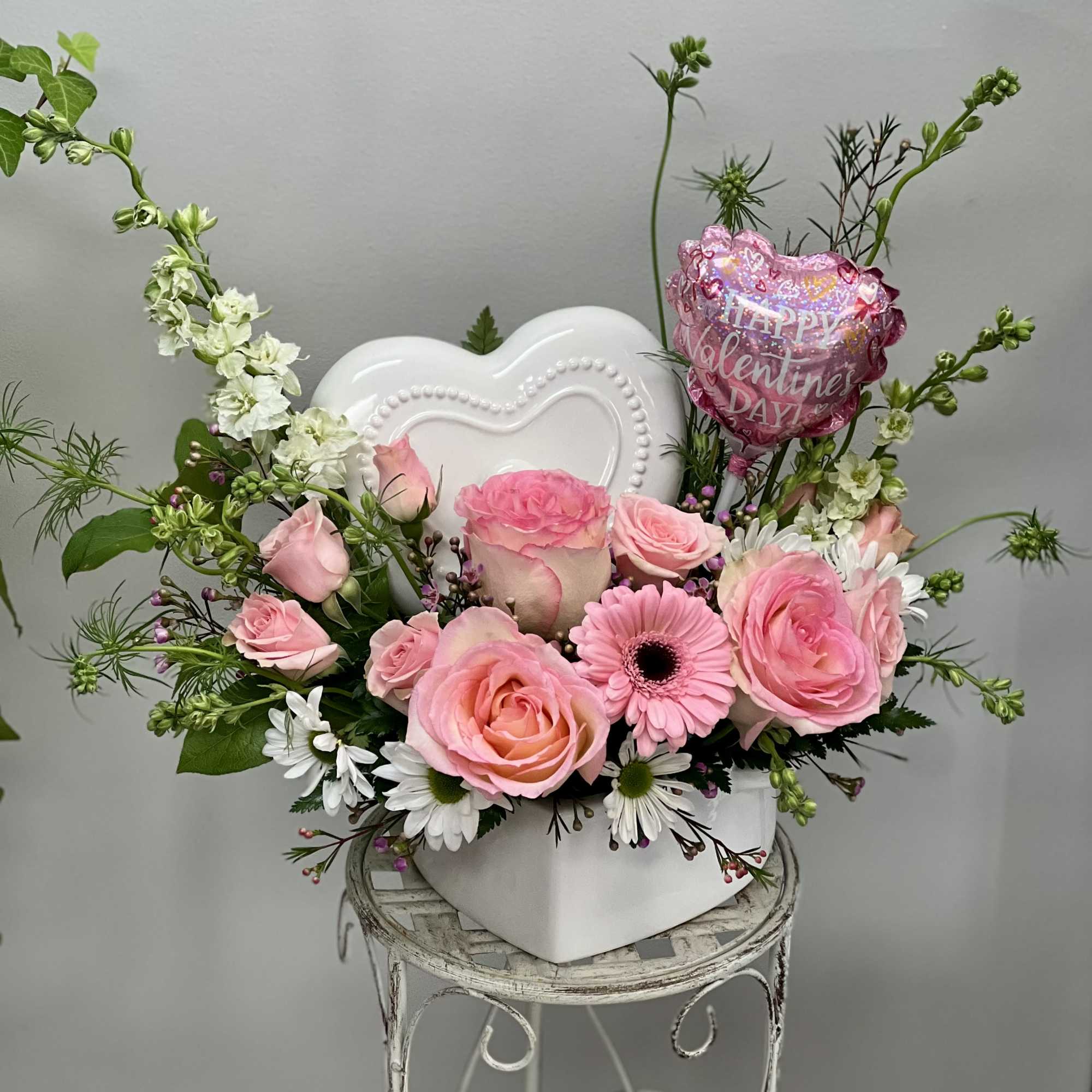 Pink roses and daisies in a white heart-shaped container with a Valentine balloon