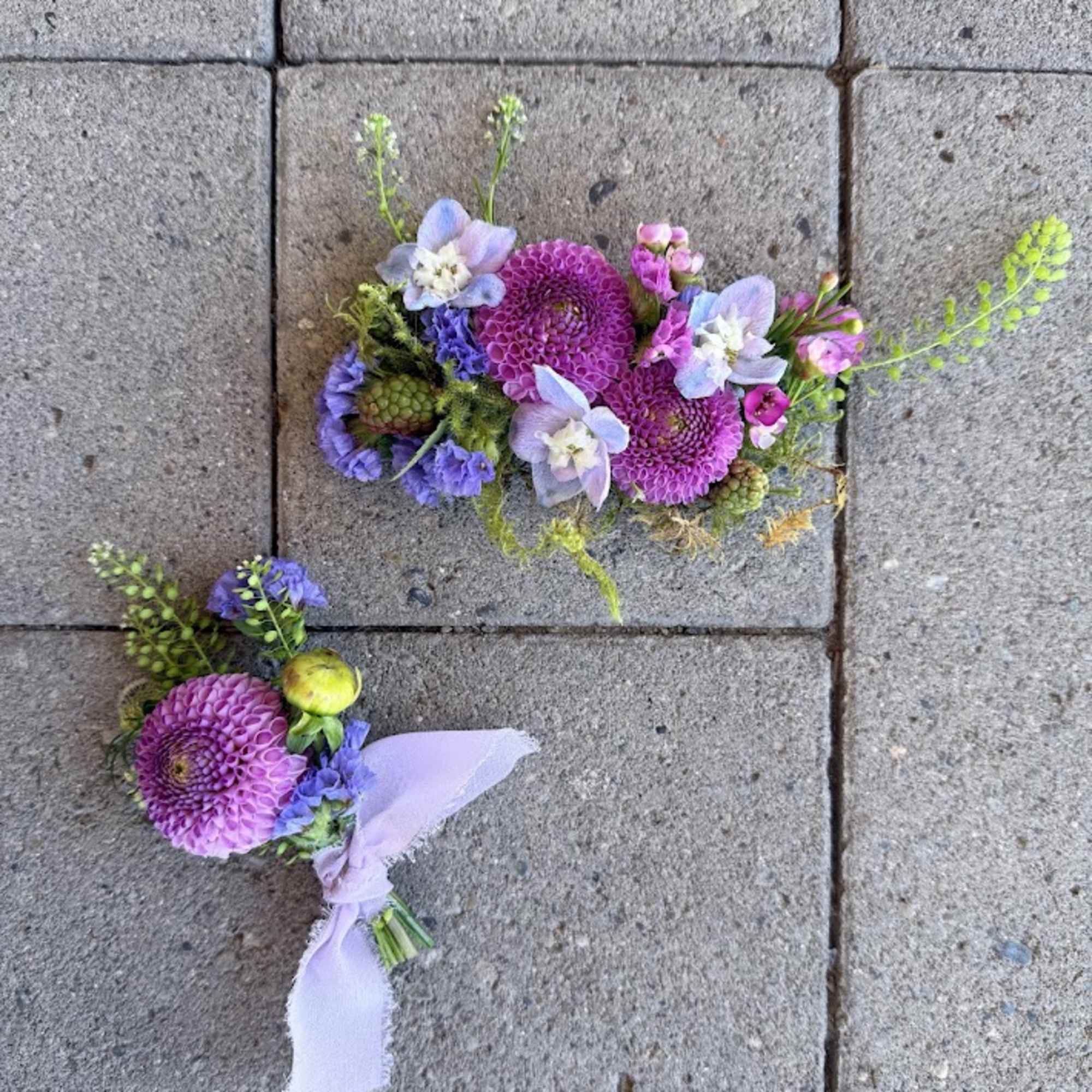 Two small purple floral boutonnieres with ribbon on pavement