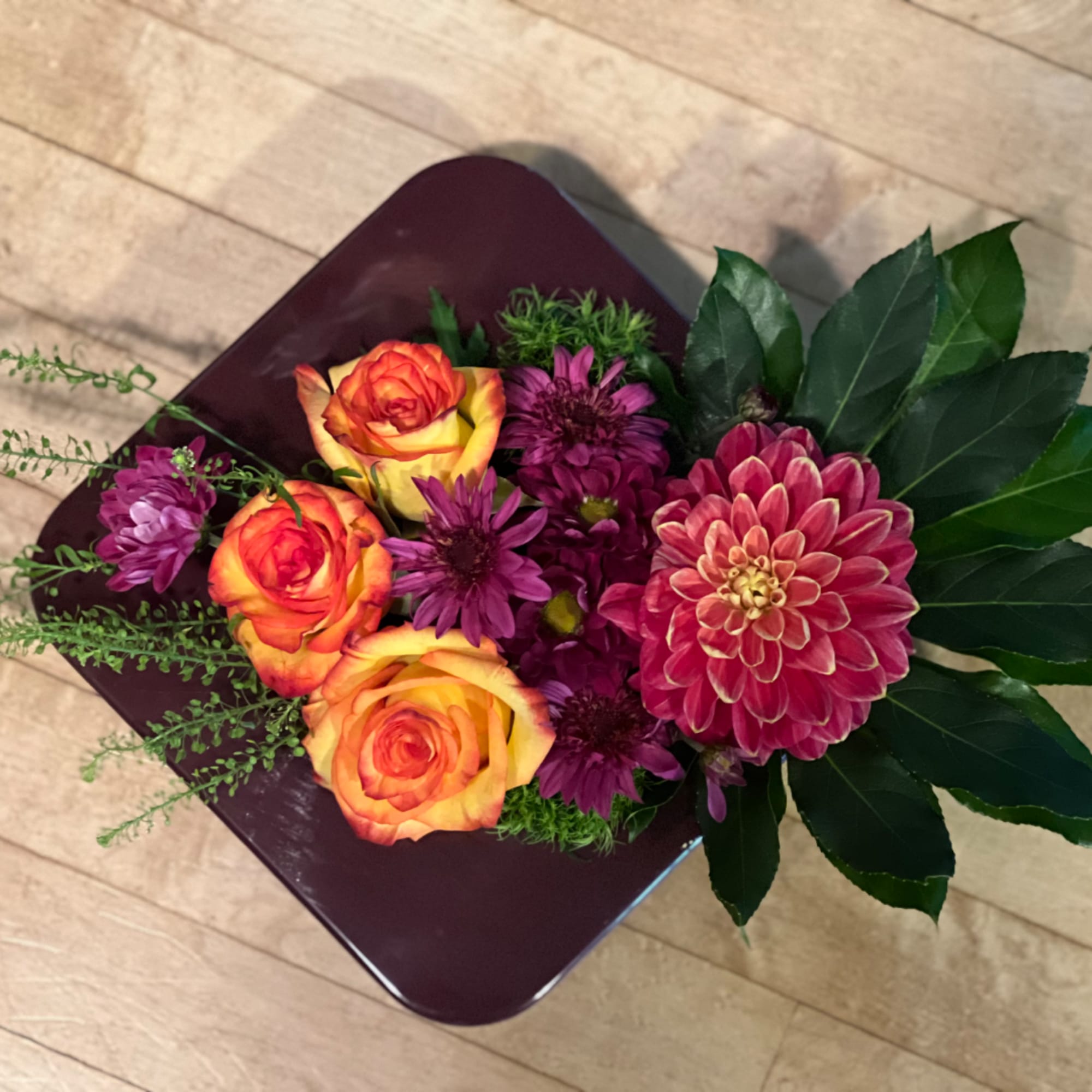 Mixed bouquet with orange-yellow roses and purple blooms in a dark square container