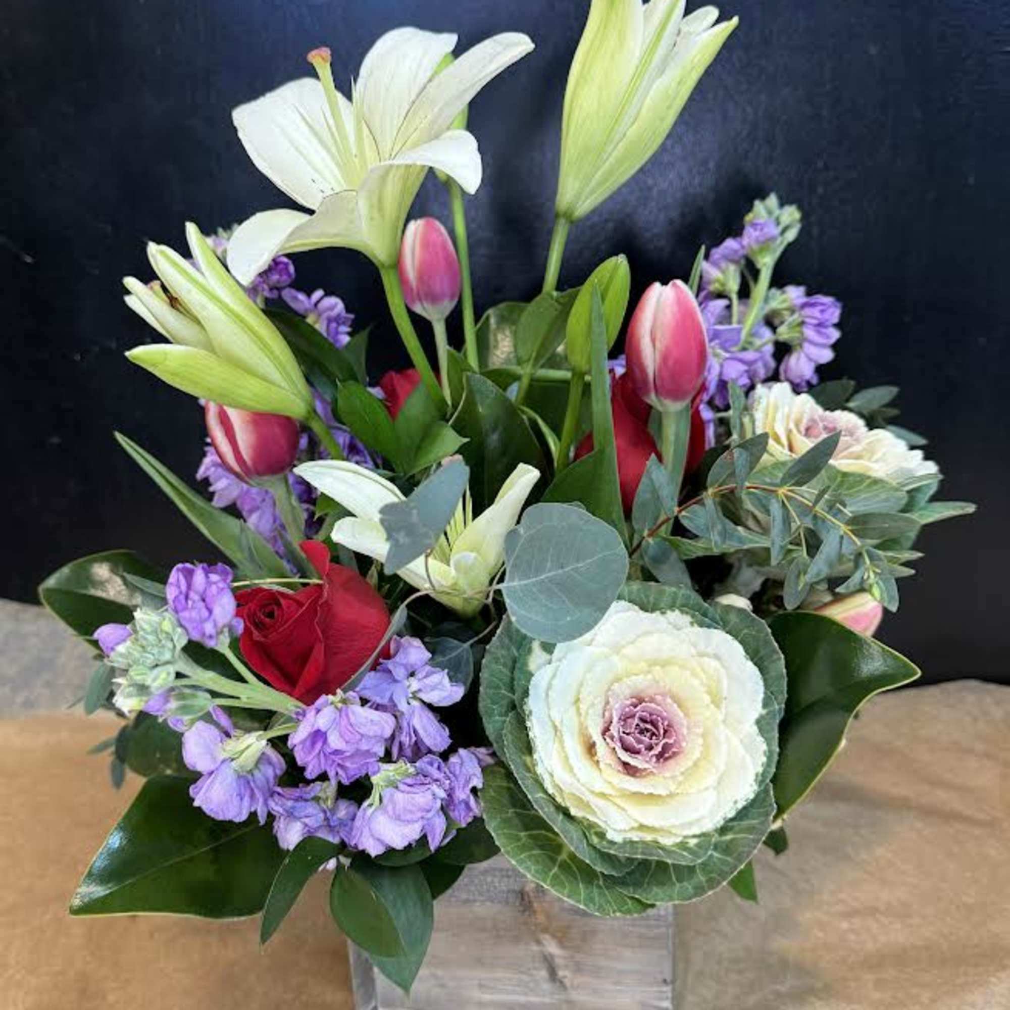 Mixed bouquet with lilies, roses, and purple blooms in a clear square vase