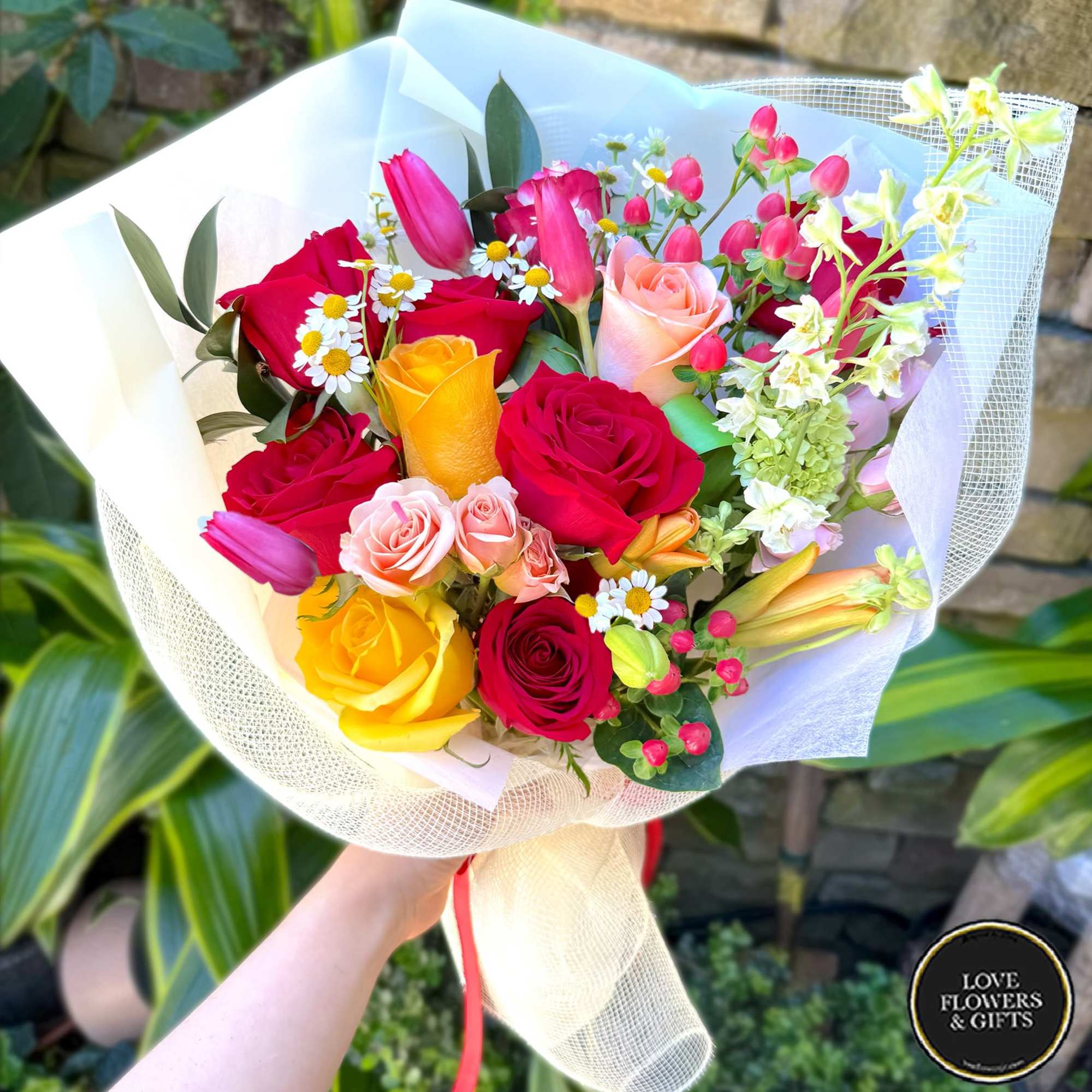 Handheld bouquet of red, yellow, and pink flowers wrapped in white paper and mesh.