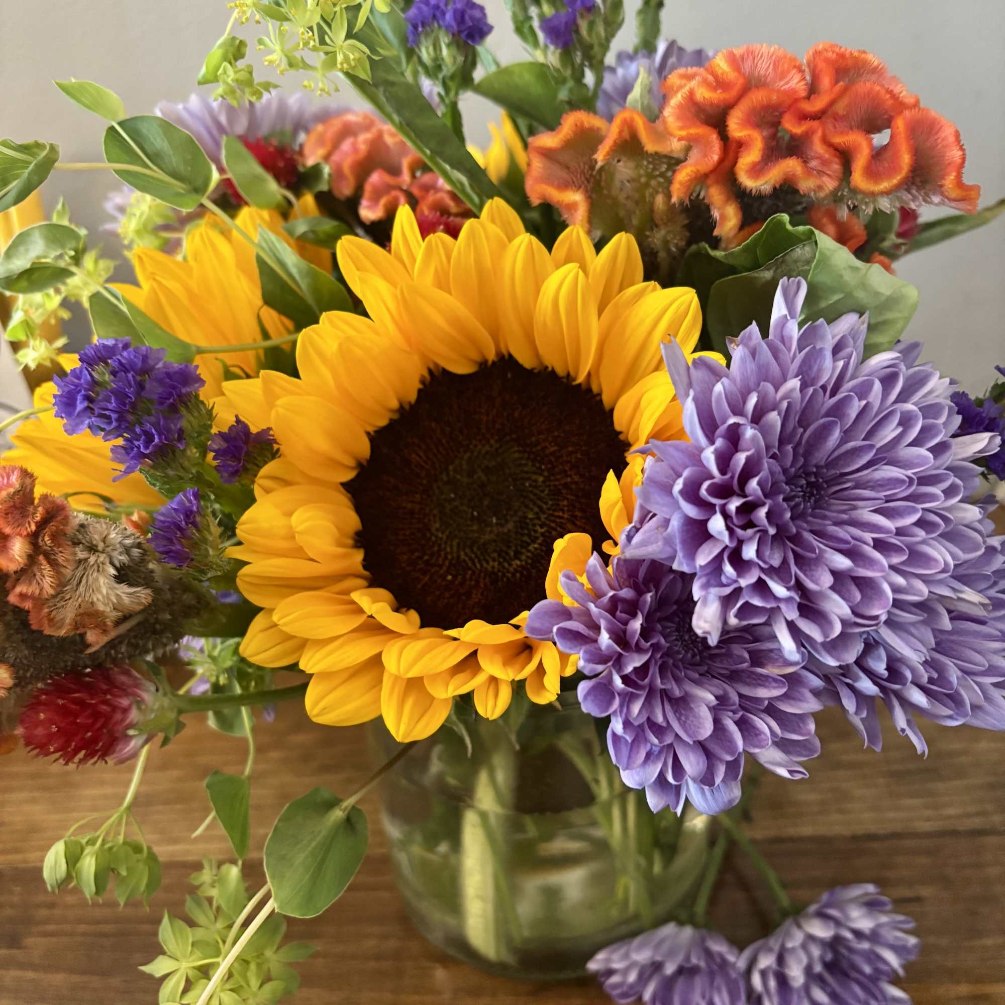 Mixed bouquet with a sunflower, purple chrysanthemums, and orange celosia in a glass vase