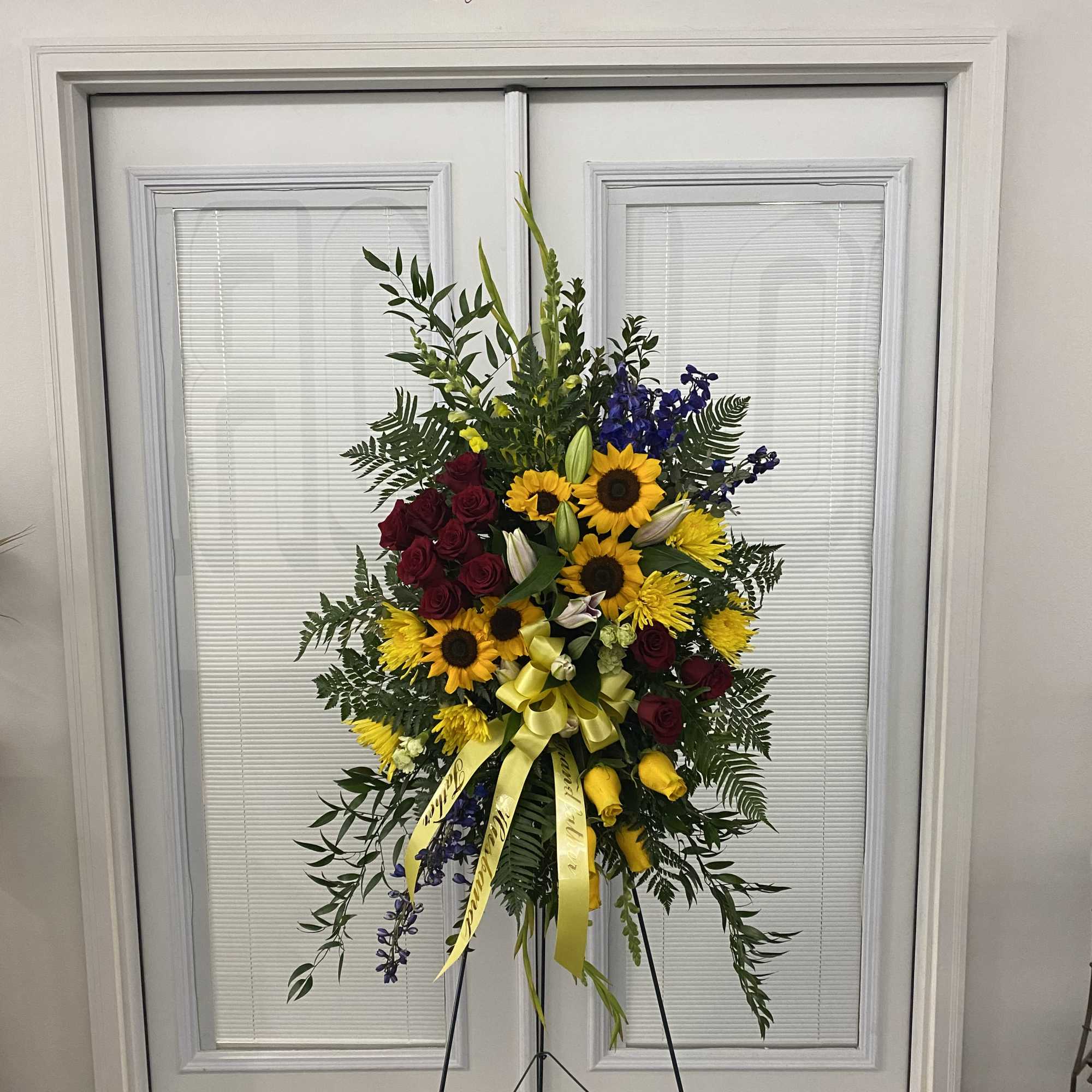 Standing floral spray with red roses, yellow sunflowers, and blue flowers on an easel