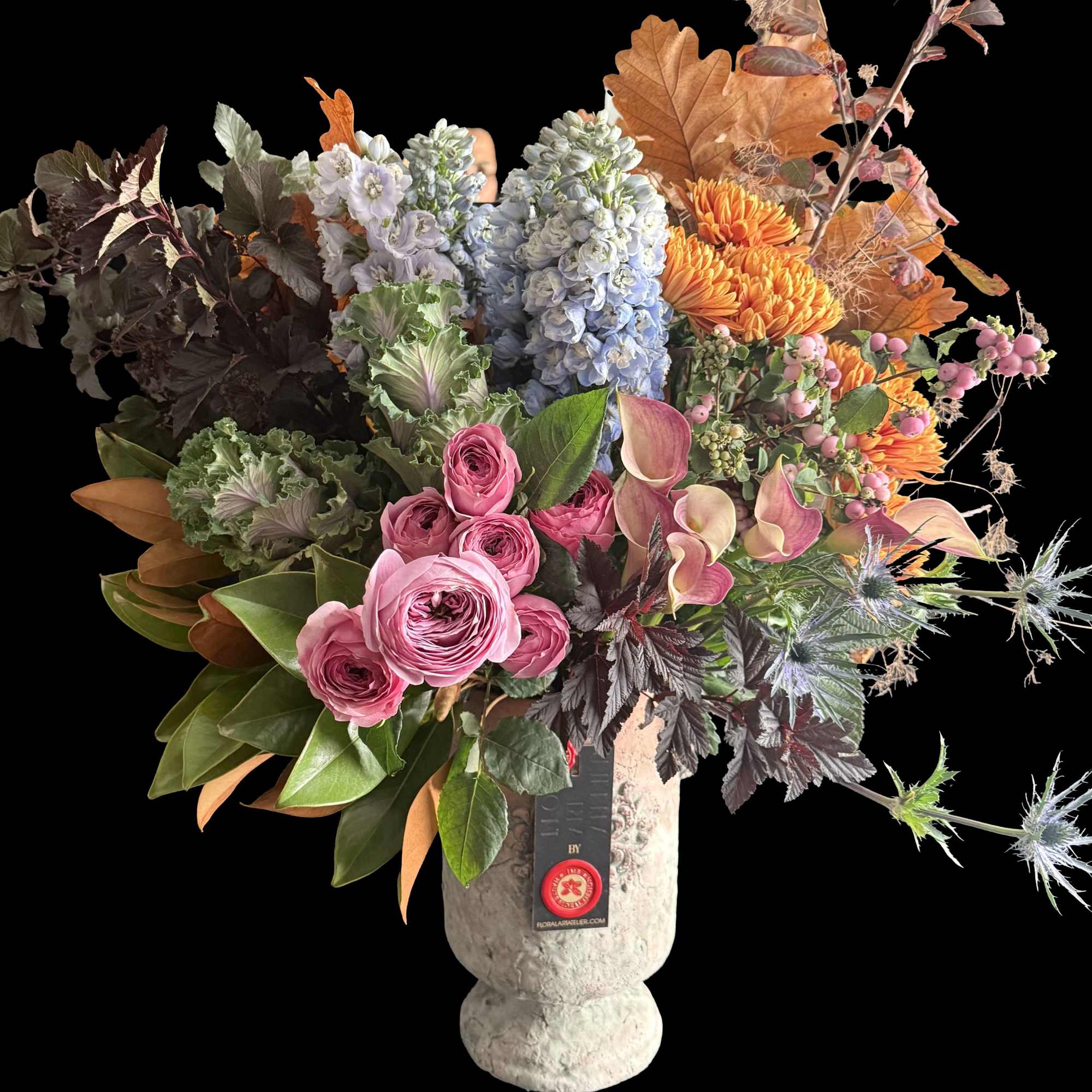 Tall mixed arrangement with pink ranunculus, orange mums, blue delphinium, and calla lilies in a stone urn vase