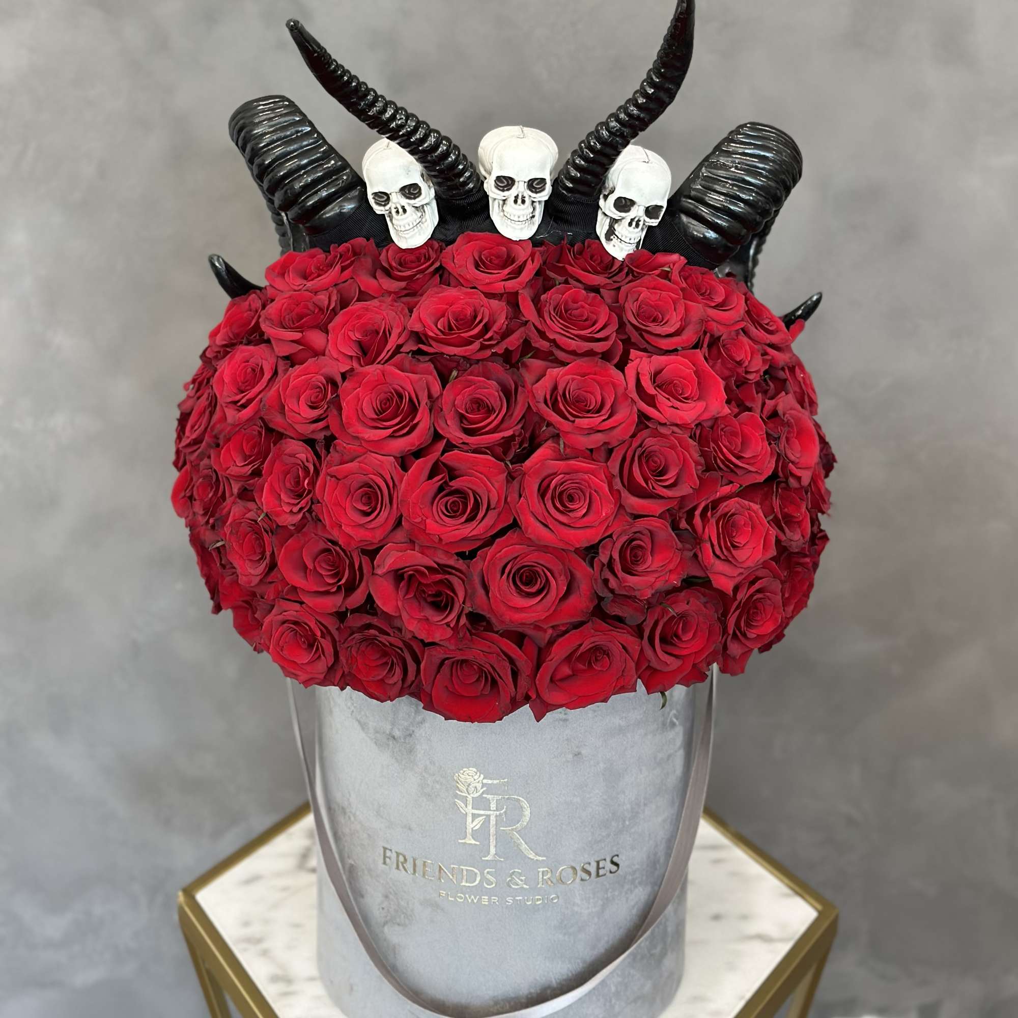 Red roses arranged in a gray hat box with black horn decorations and skull toppers
