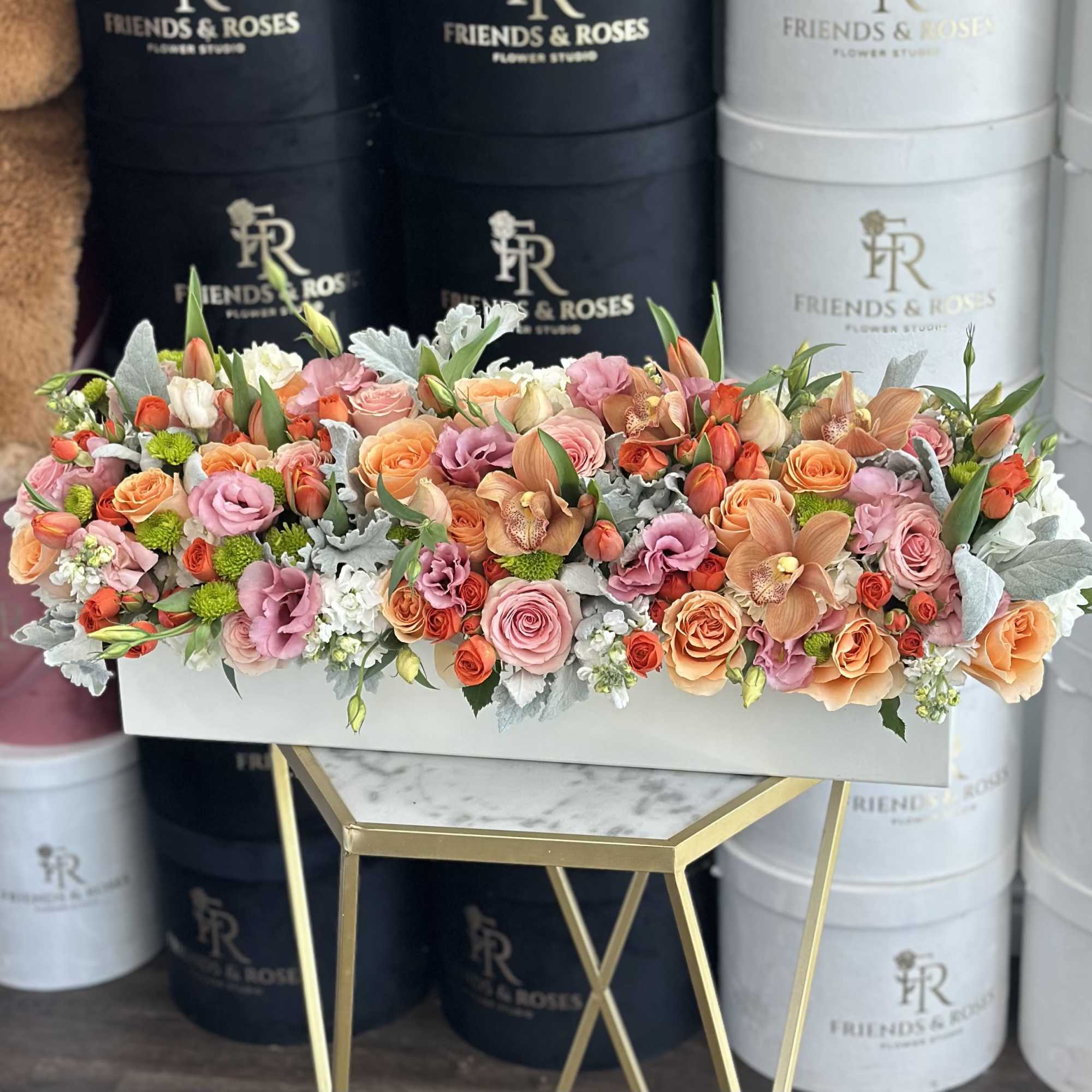 Low rectangular arrangement of peach, orange, and pink roses, tulips, and orchids in a white box.