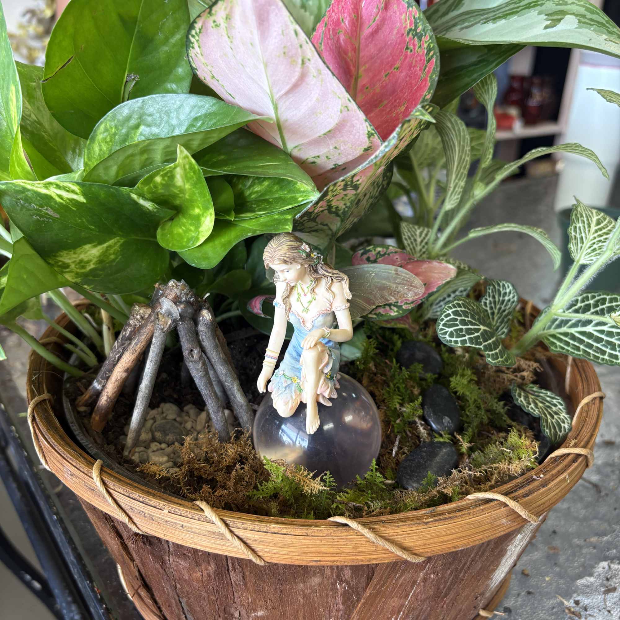 Potted tropical plant arrangement in a wooden basket with a fairy figurine