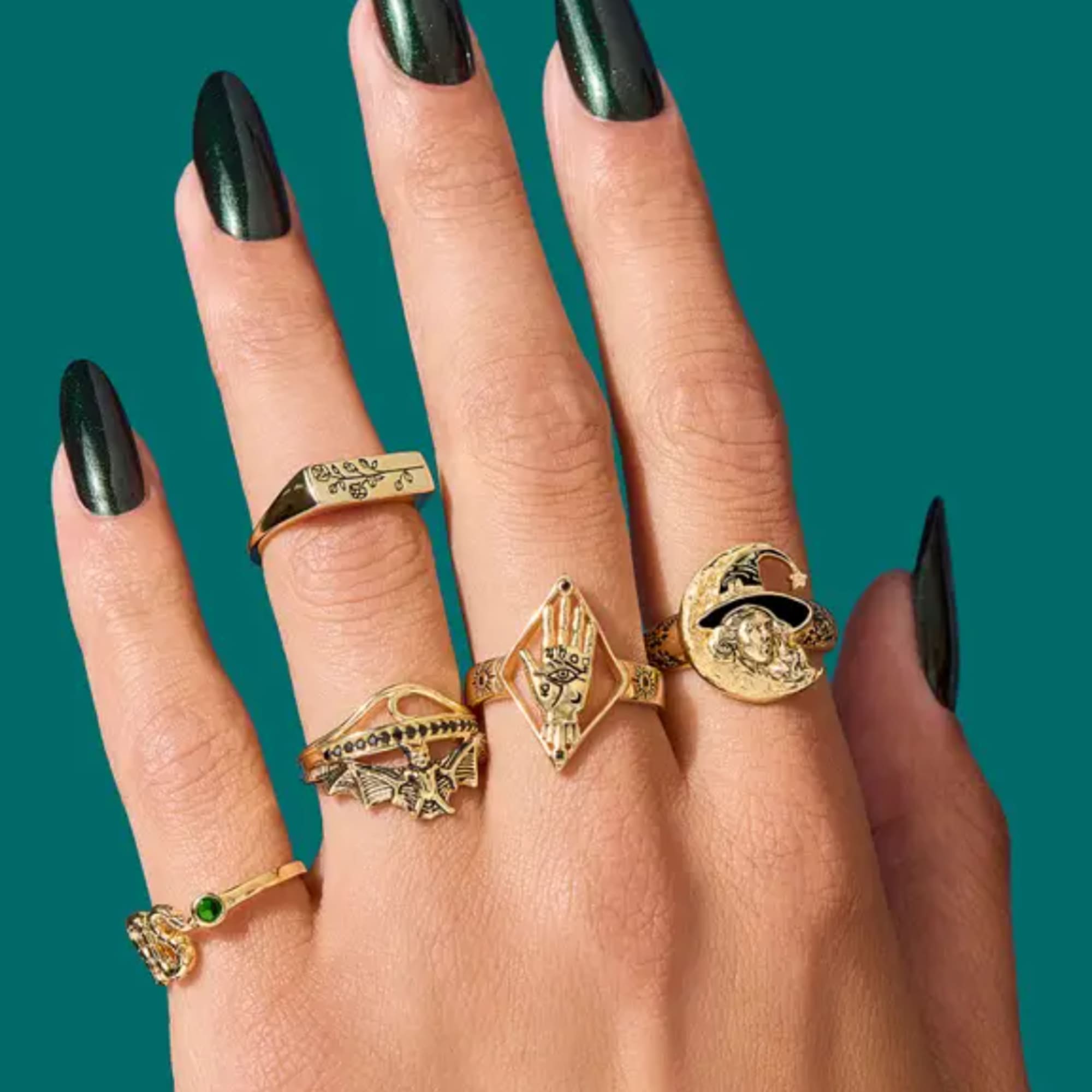 Hand wearing several gold rings with black nails on a teal background