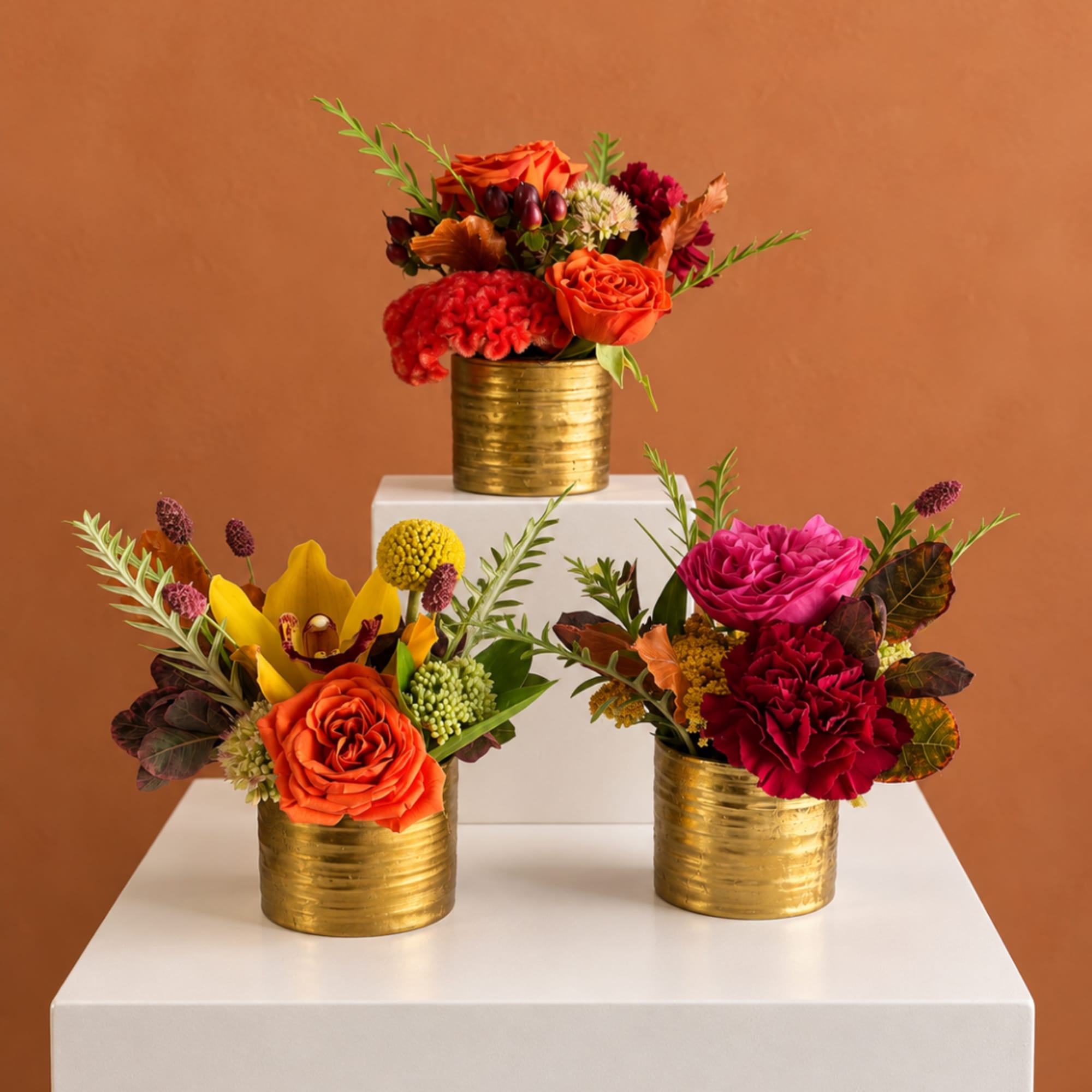 Three floral arrangements in gold containers on white pedestals