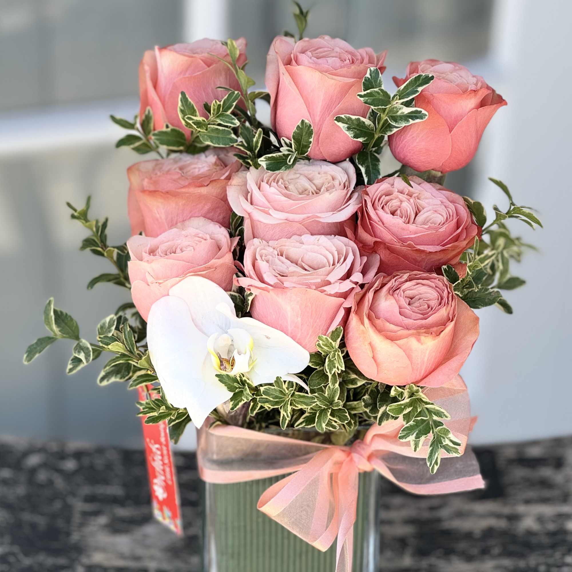 Pink roses arranged in a clear glass vase with a white orchid and ribbon