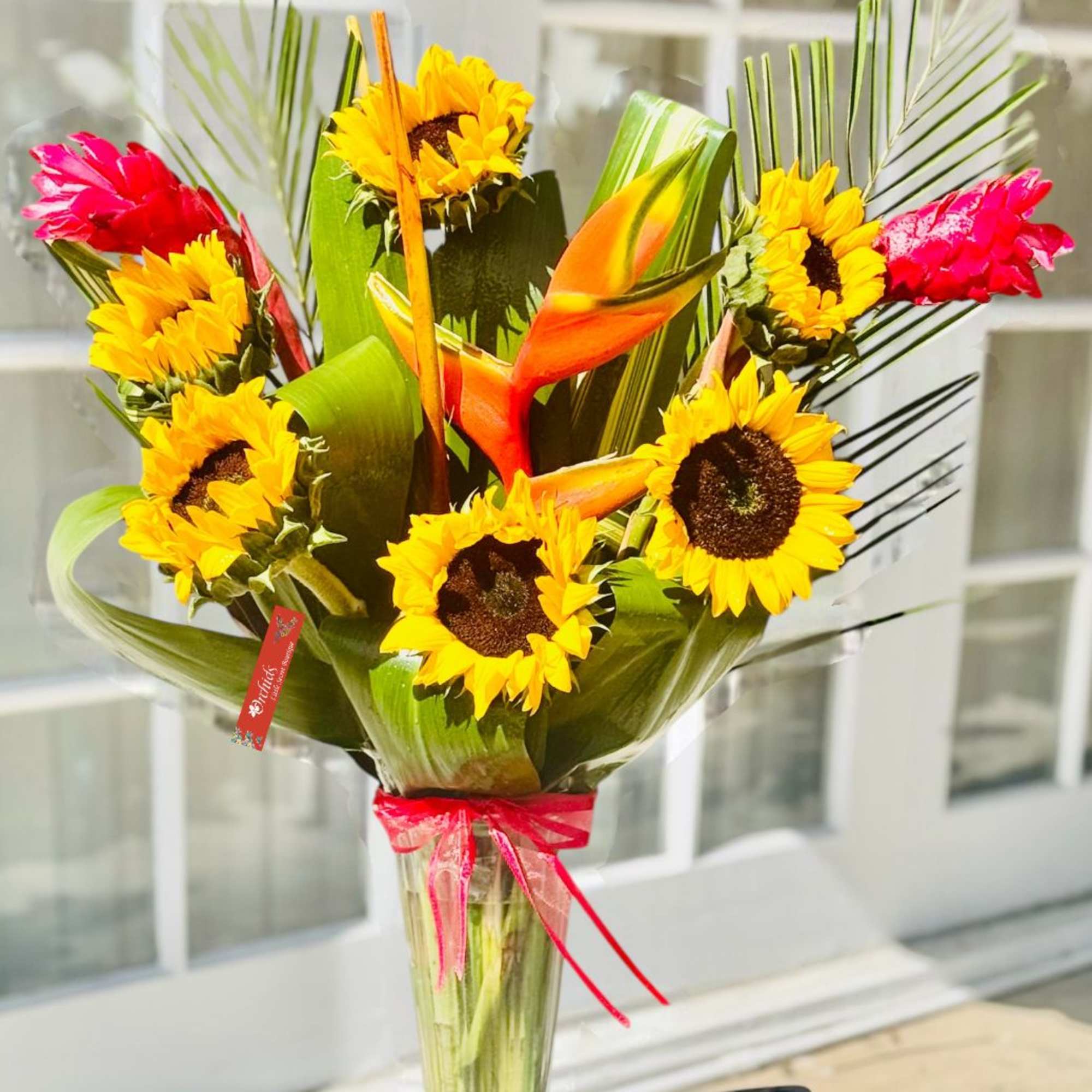 Sunflowers and tropical flowers in a clear glass vase with a pink ribbon